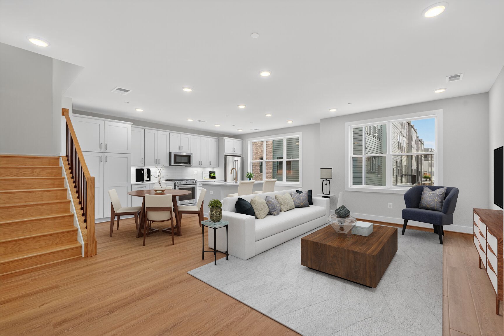 A modern living and dining area with a white sofa and coffee table in the foreground, and a kitchen with white cabinets and stainless steel appliances in the background.
