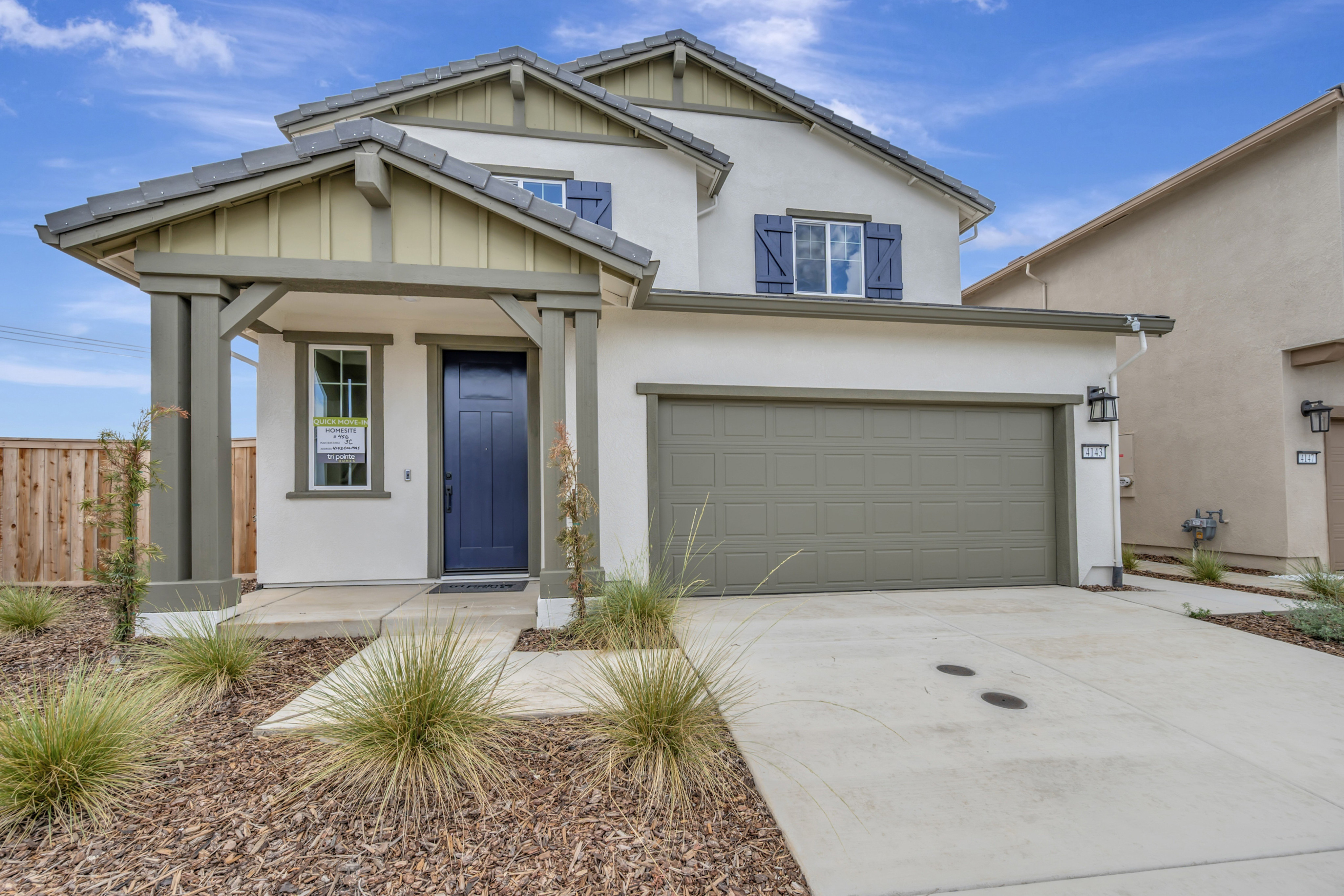 A modern stucco house with a dark blue door and olive green garage door stands behind a walkway bordered by ornamental grasses and wood chips, under a bright blue sky with scattered clouds.
