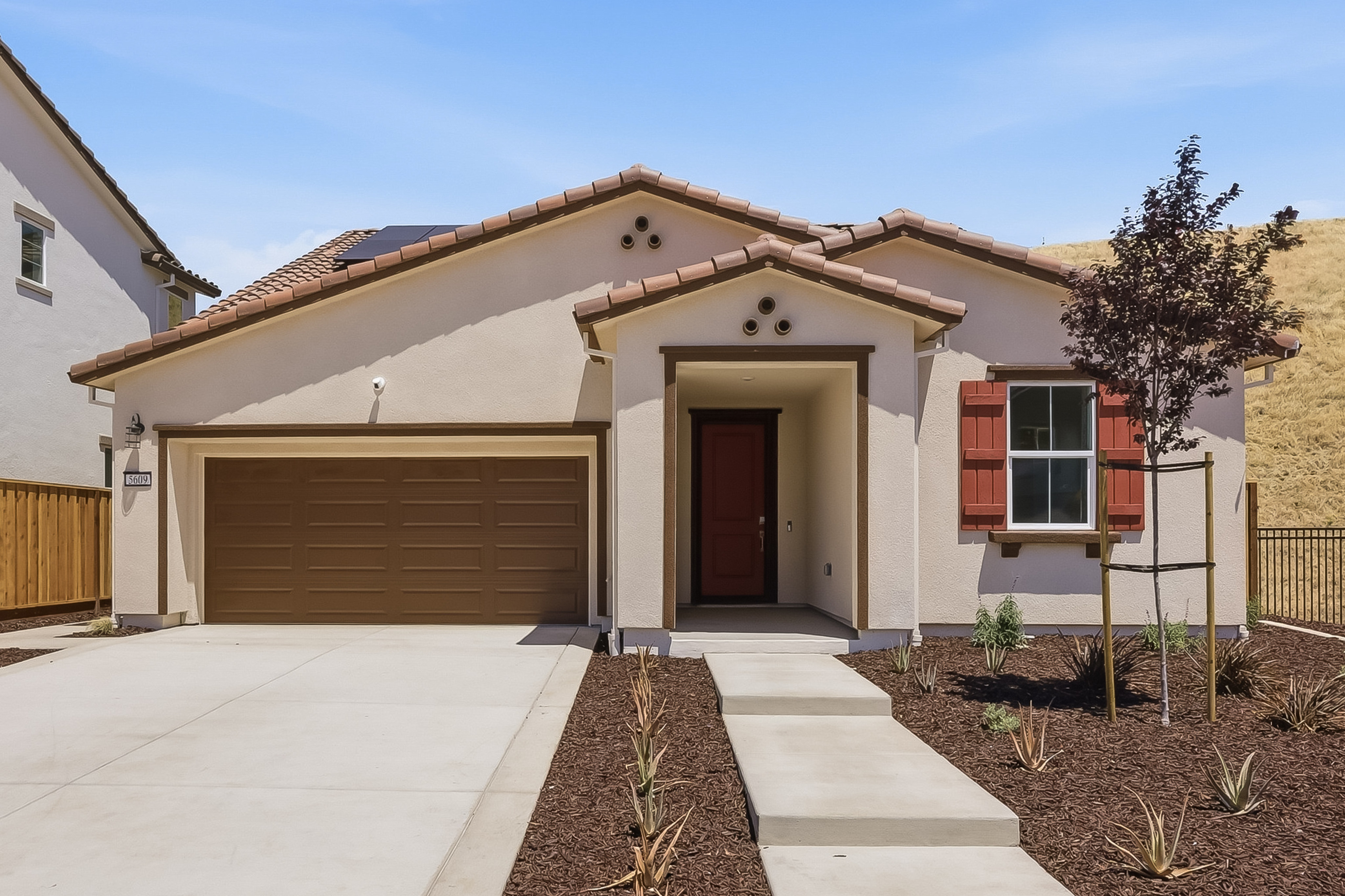 A modern stucco house with a brown garage door and red shutters sits on a concrete driveway, backed by a rolling, grassy hillside under a clear blue sky.