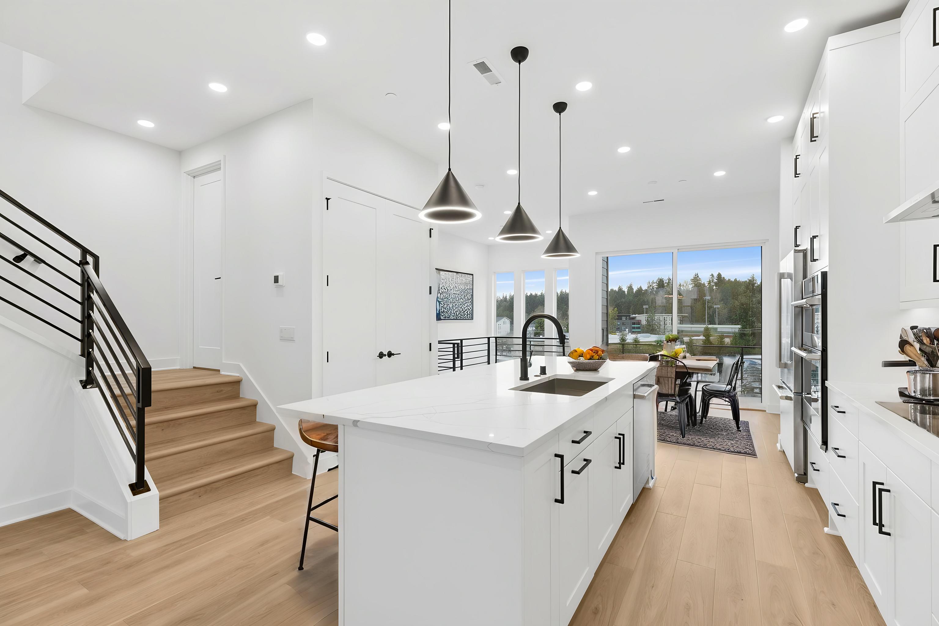 A modern white kitchen island with pendant lights in the foreground leads to a dining area and a scenic view of trees and water in the background, with a staircase to the left.