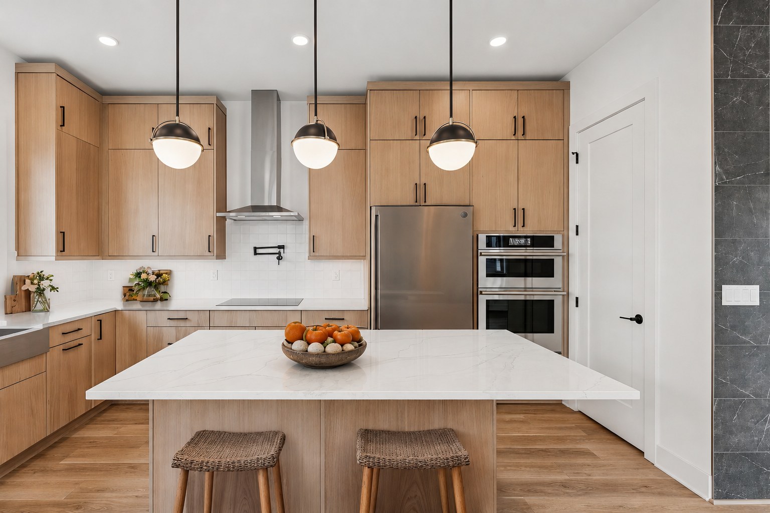 A modern kitchen with a white island and stools in the foreground, and light wood cabinets and stainless steel appliances in the background.