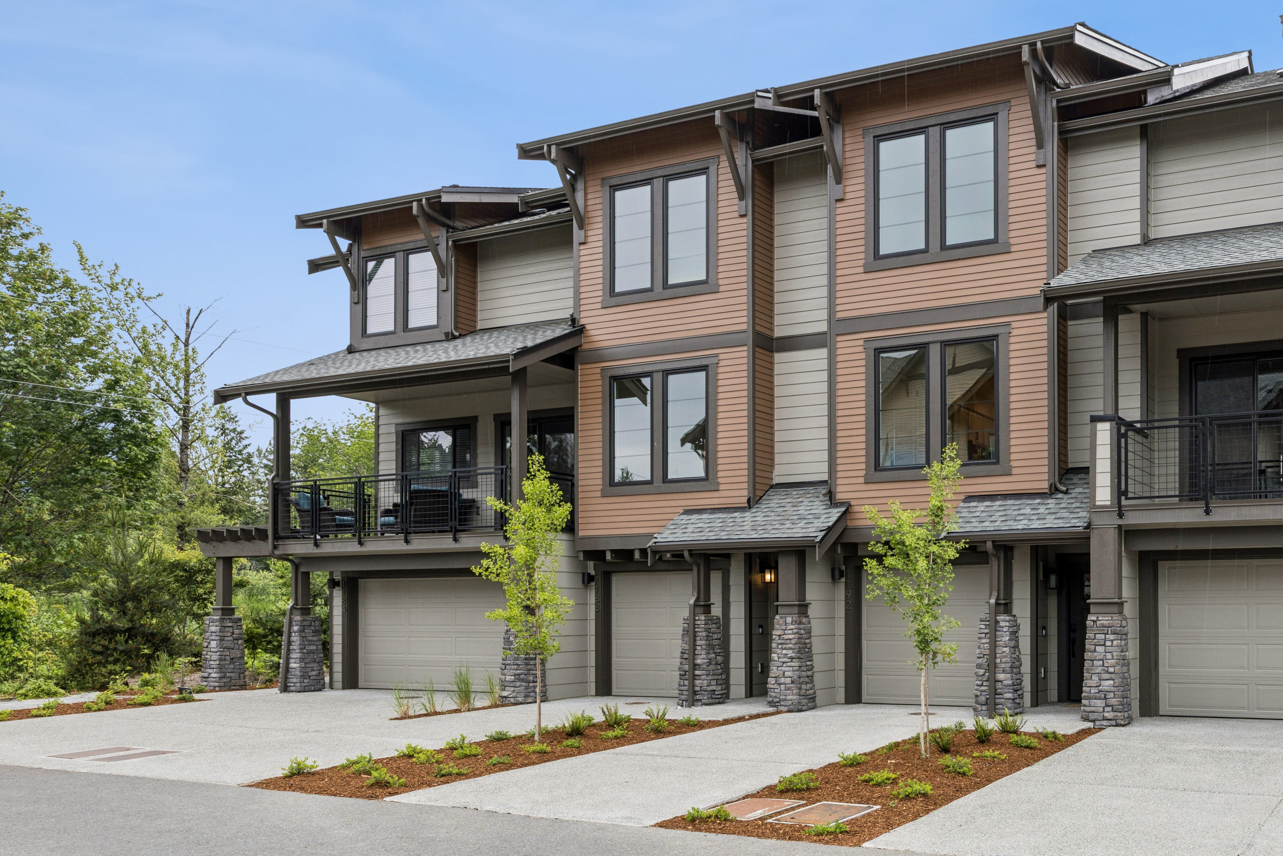 Modern townhouses with attached garages and manicured landscaping are set against a backdrop of lush green trees under a clear sky.