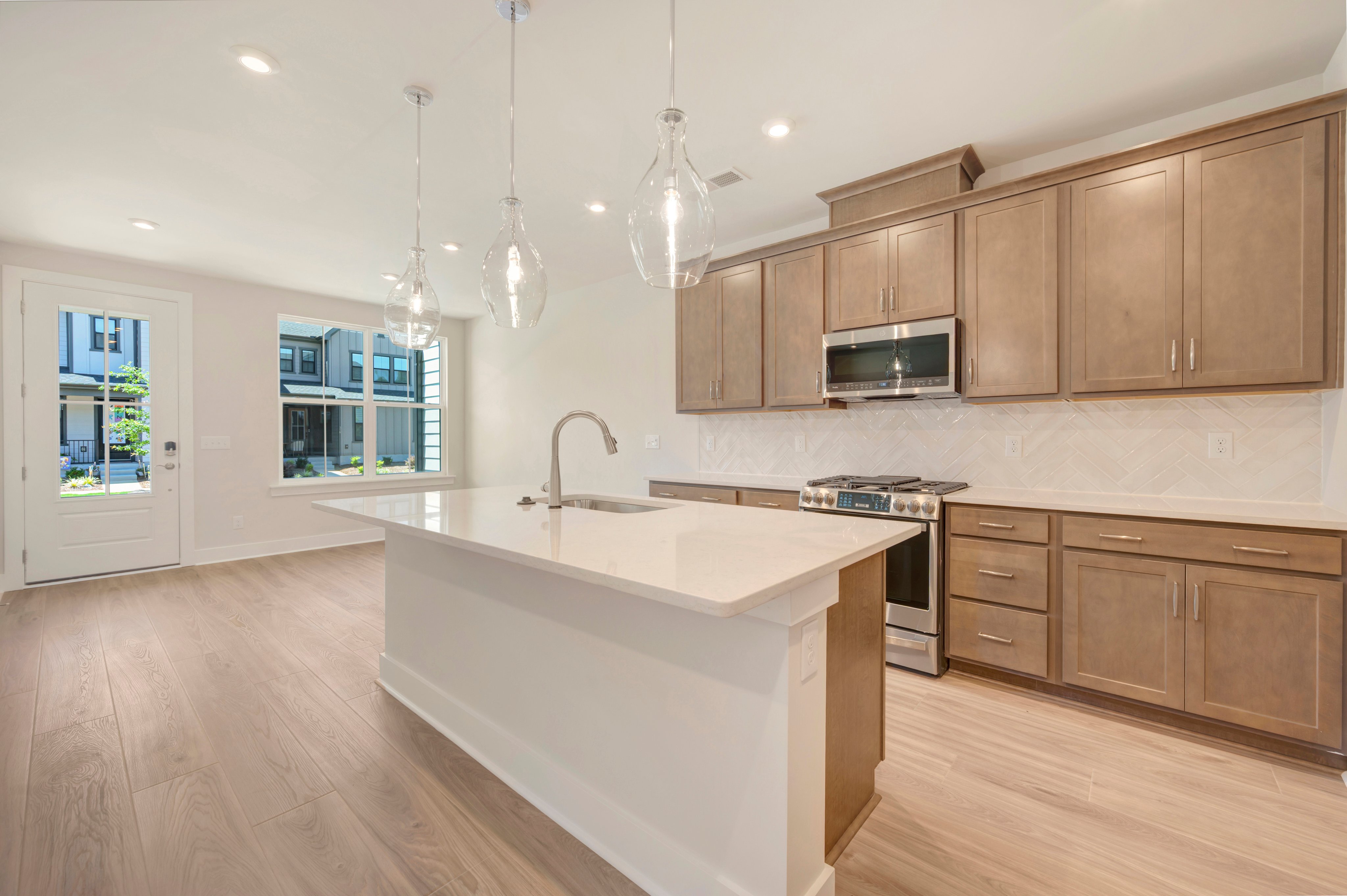 A modern kitchen island with a sink is in the foreground, while a row of wooden cabinets and a stainless steel stove are in the background, with a large window showing an exterior view.