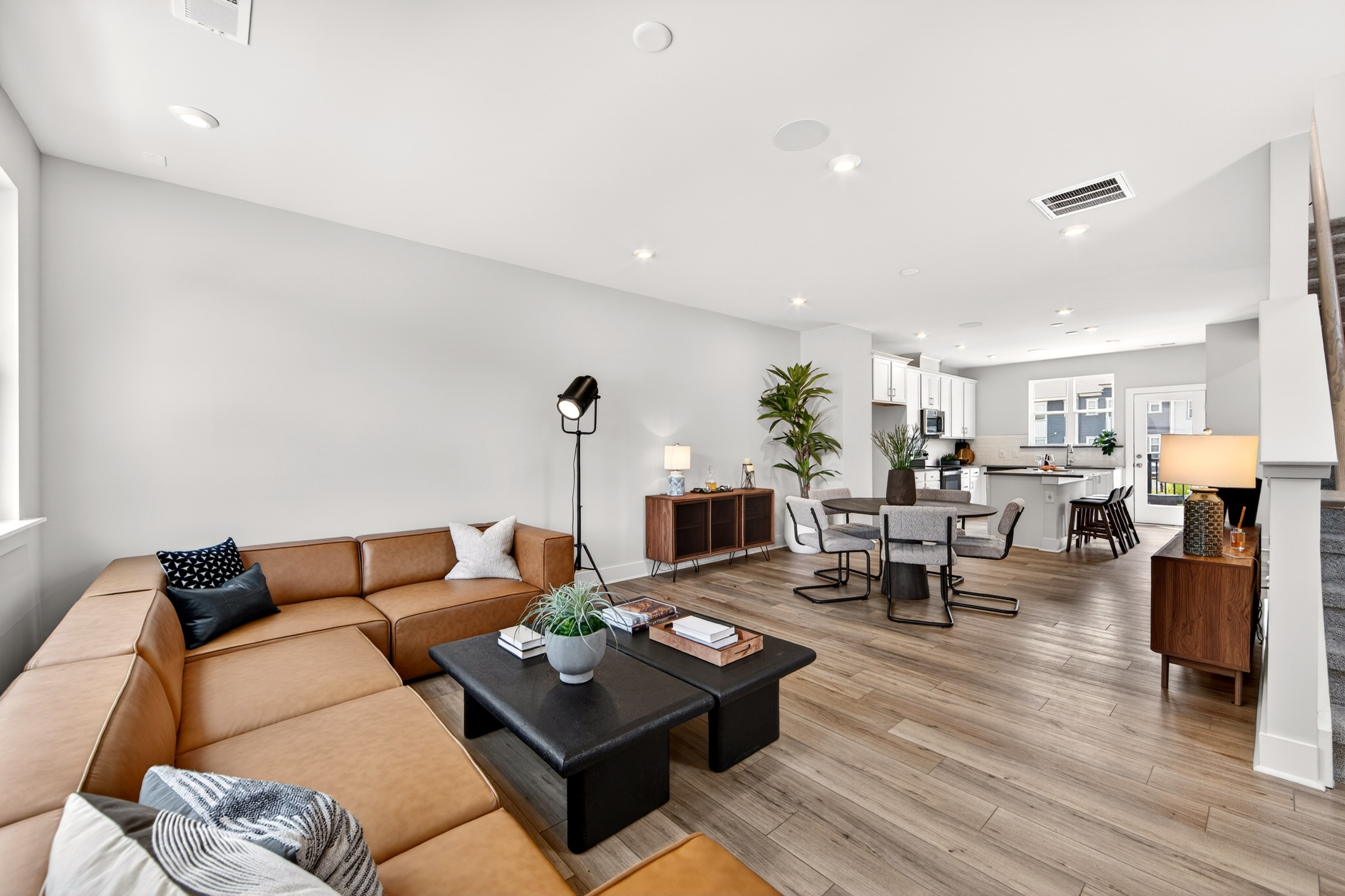 A spacious, modern living room with a large sectional sofa and coffee table in the foreground opens to a dining area and kitchen in the background.