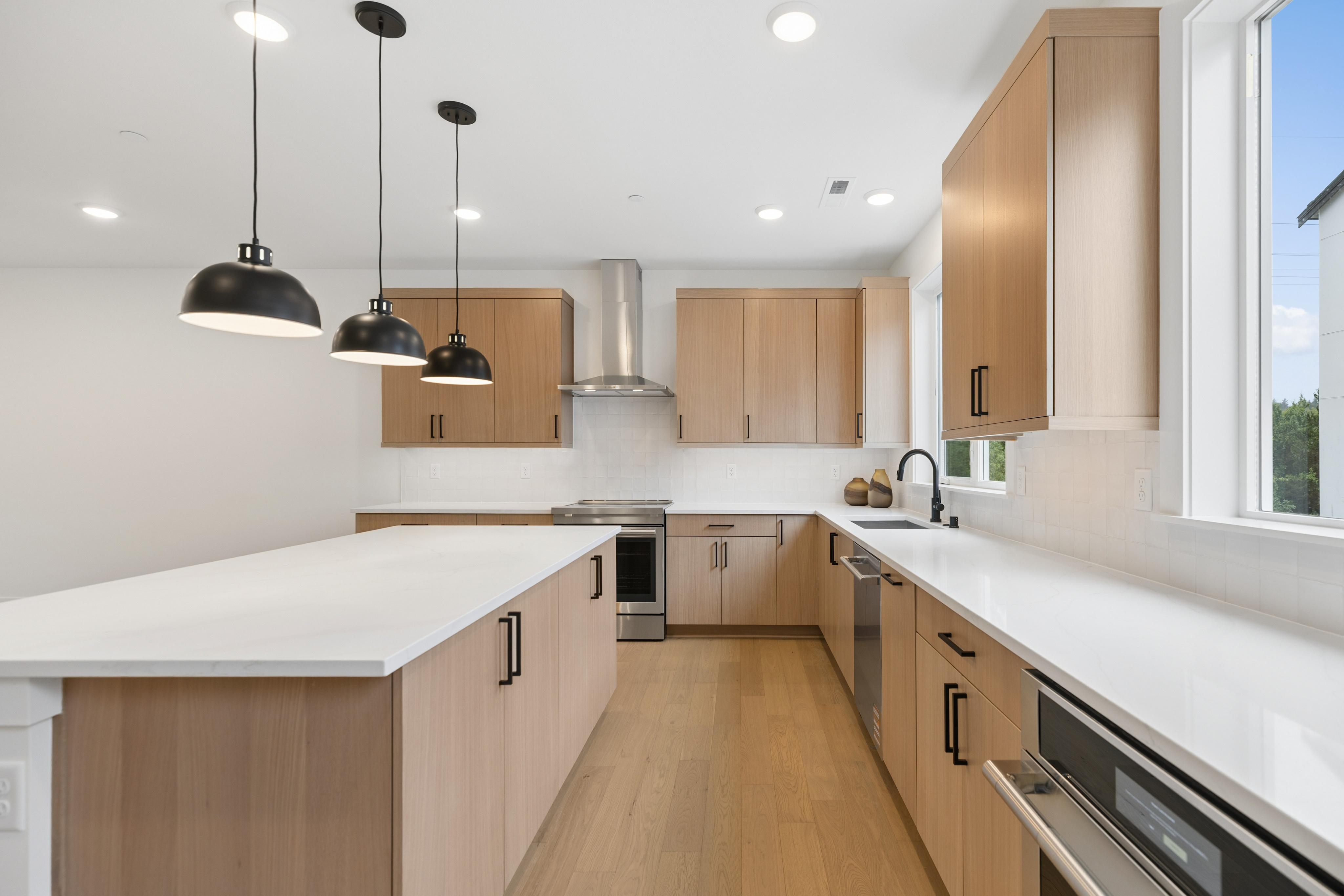A modern kitchen with a large island in the foreground and light wood cabinets and stainless steel appliances in the background.