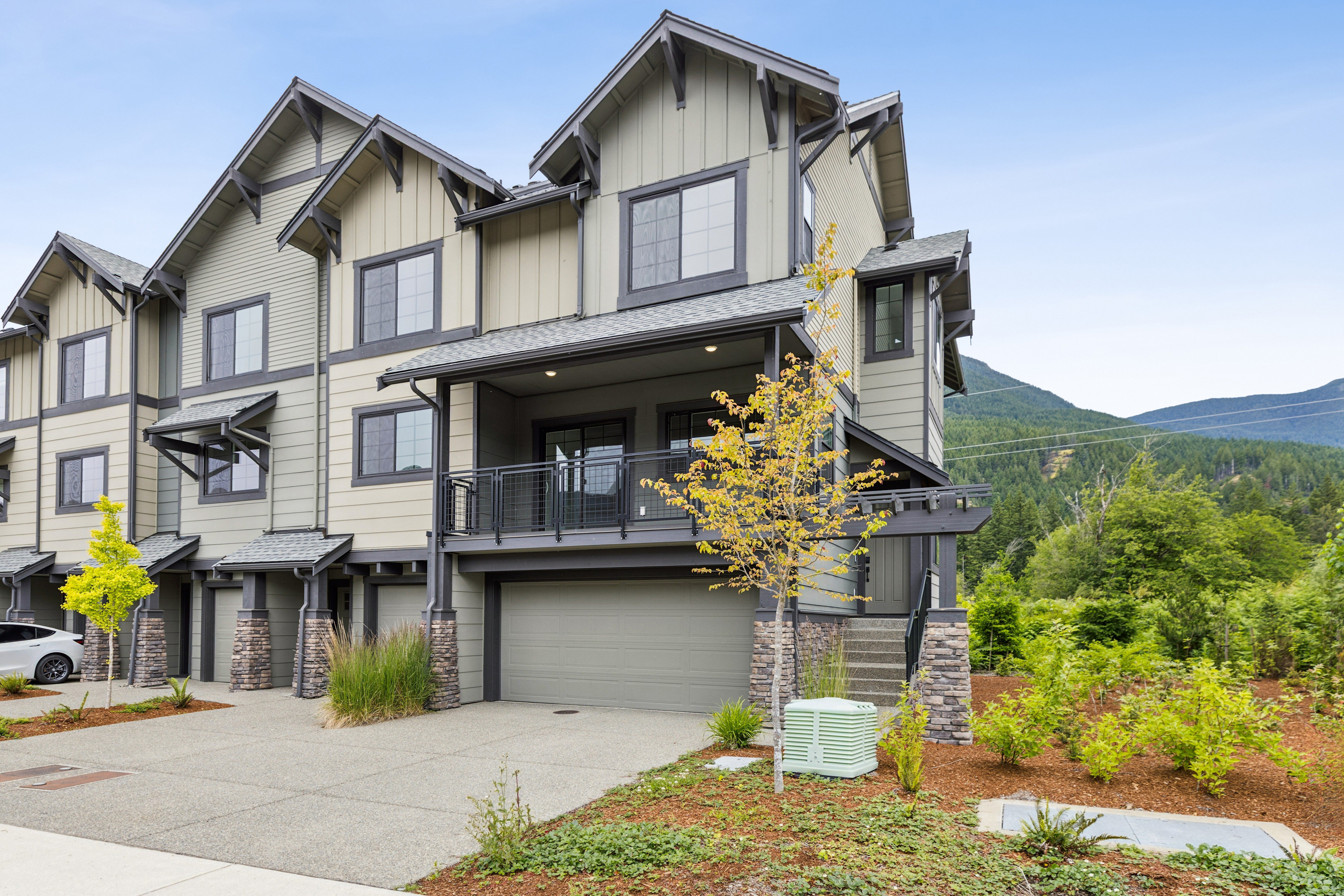 A row of modern townhouses with garages in the foreground sits against a backdrop of lush green trees and distant mountains.