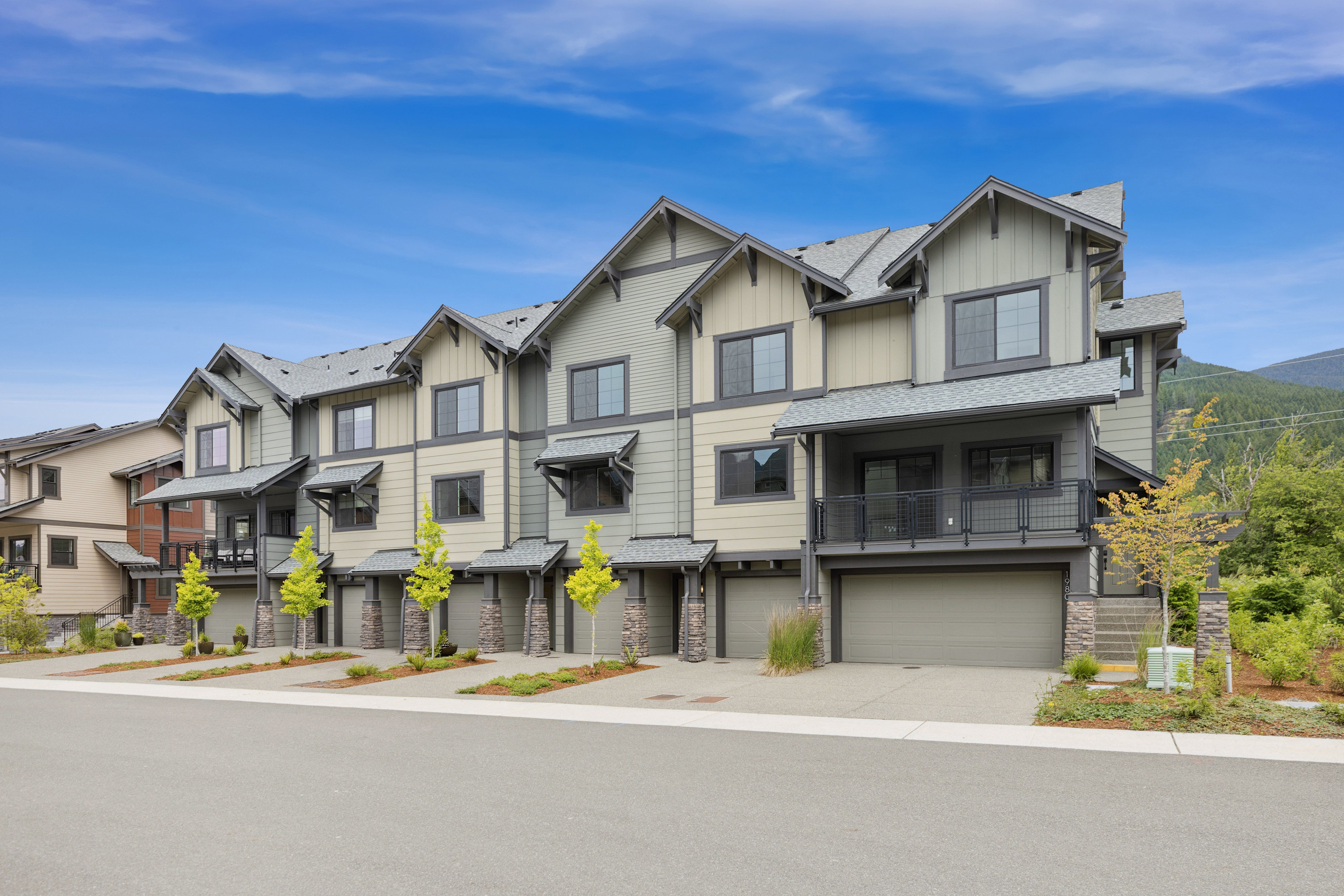 A row of modern townhouses with attached garages sits under a bright blue sky with distant mountains.