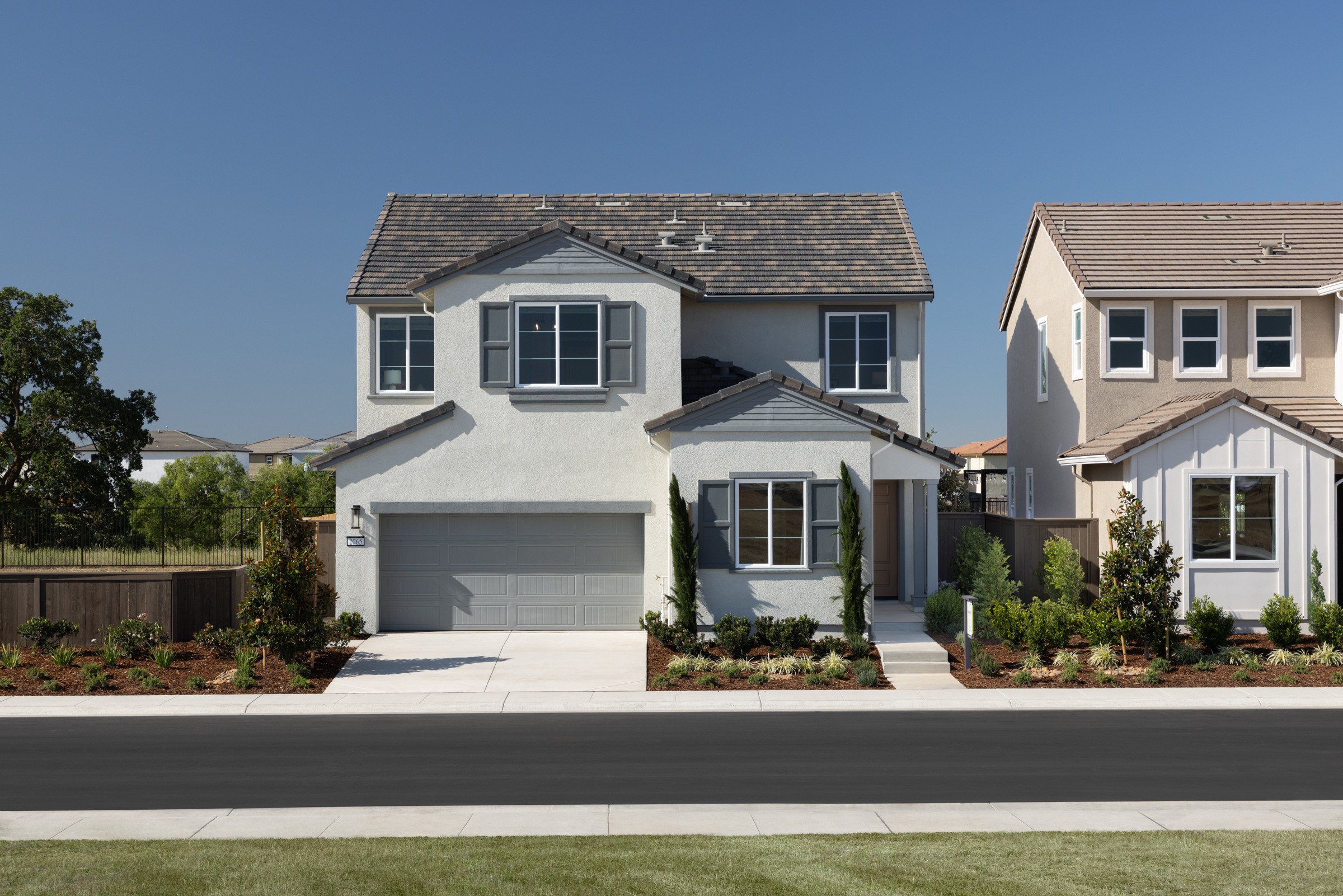 A modern two-story house with gray shutters and a garage door sits in the foreground, while other houses and trees are visible in the background under a clear blue sky.
