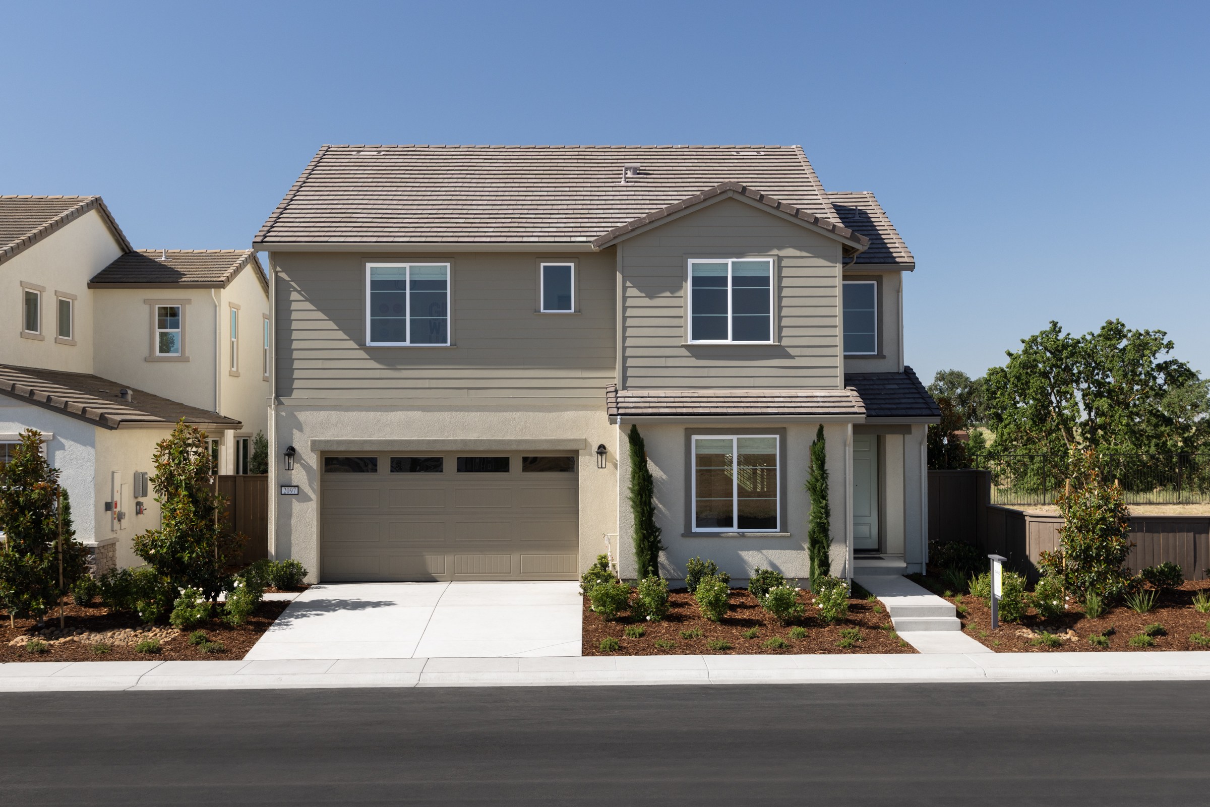 A modern two-story house with a gray exterior and a two-car garage sits on a manicured lawn, with neighboring houses and trees visible in the background under a clear blue sky.