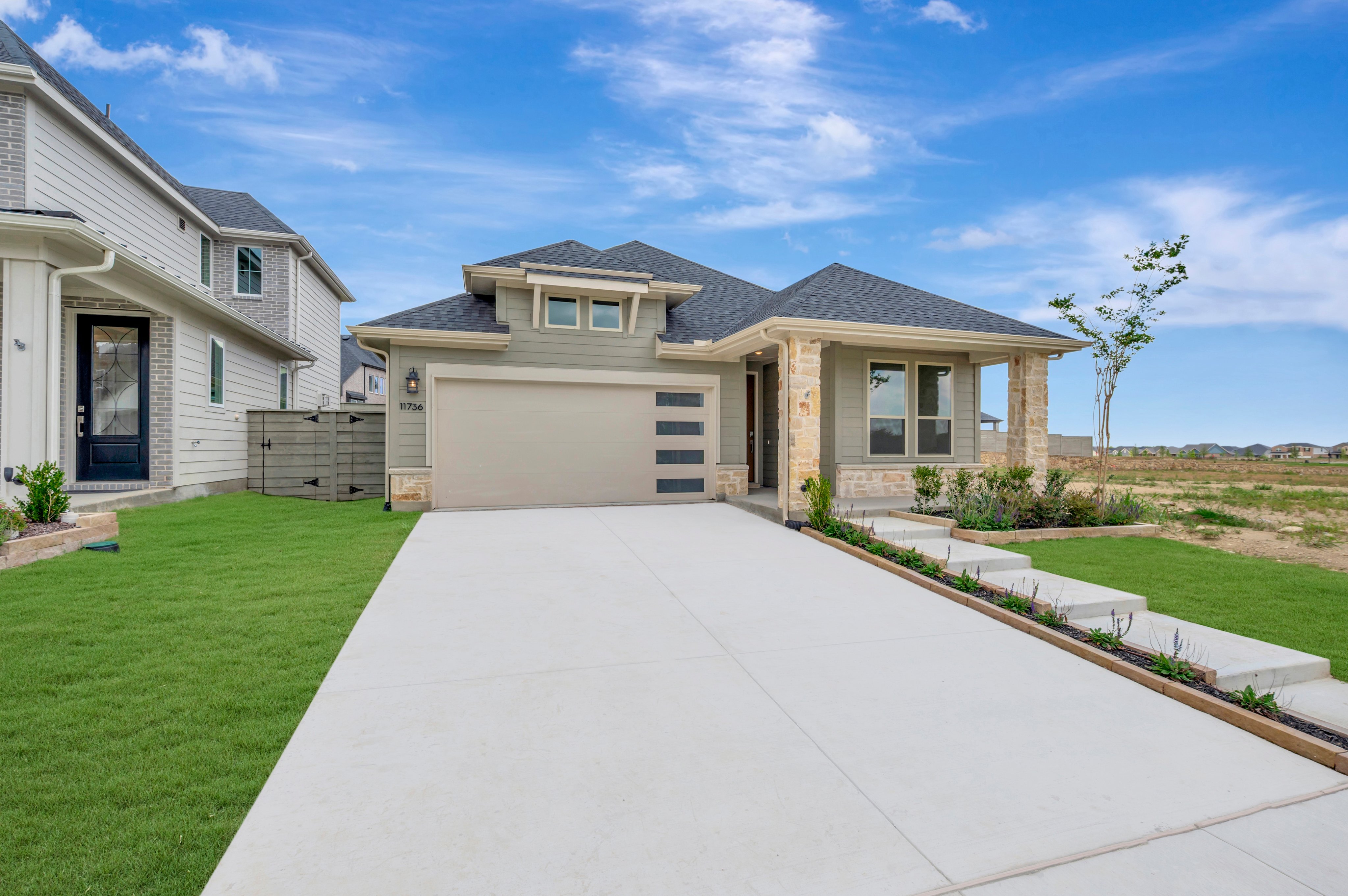 A modern, light green house with a stone-accented entrance sits on a manicured lawn, with a long concrete driveway leading to a suburban neighborhood under a bright blue sky.