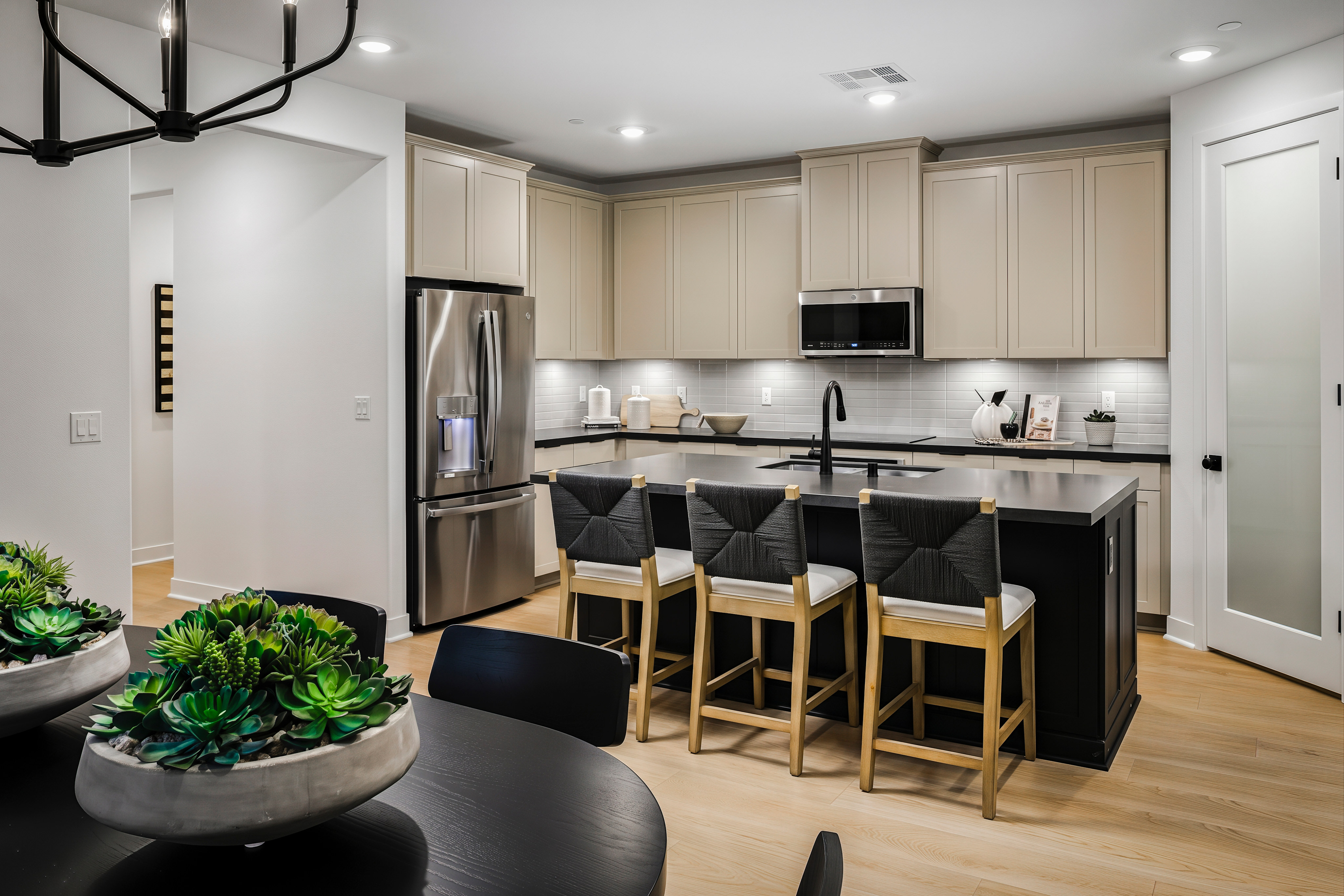 A modern kitchen with a large island and bar stools in the foreground, and a stainless steel refrigerator and light-colored cabinets in the background.