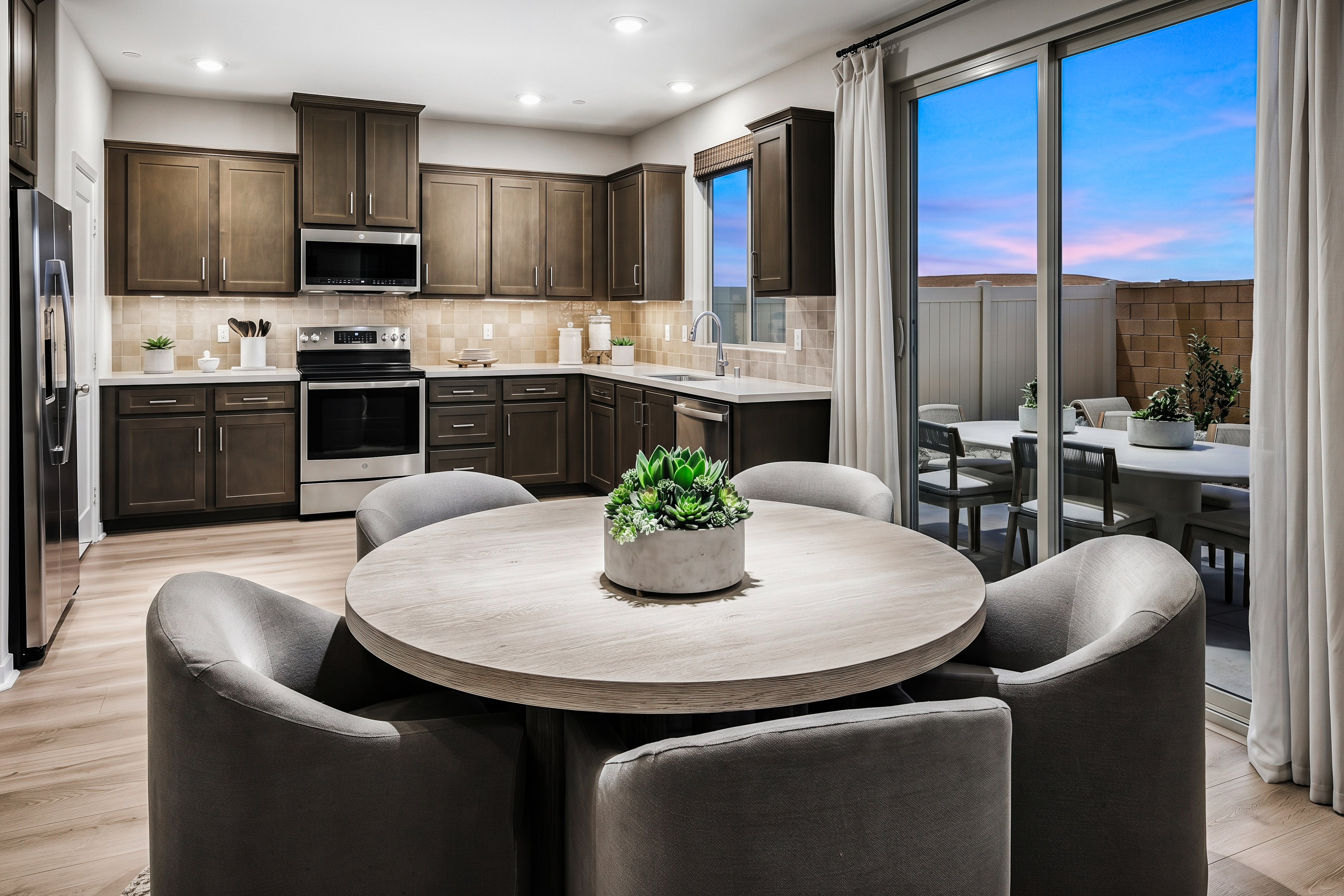 A round dining table with upholstered chairs sits in the foreground, while a modern kitchen with dark cabinetry and stainless steel appliances is visible in the background, leading to a patio with outdoor seating.