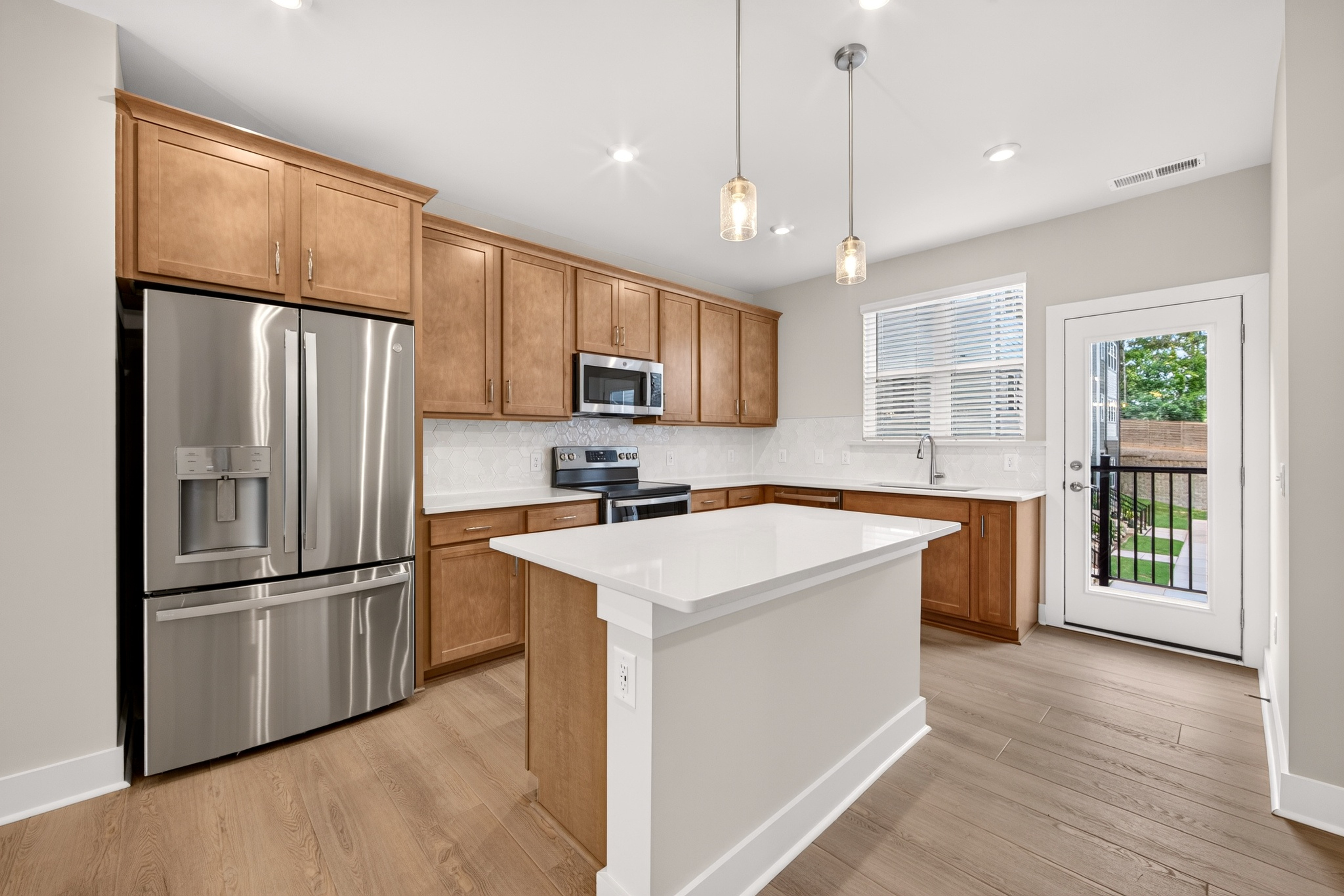 A modern kitchen with a stainless steel refrigerator and island in the foreground, and a door leading to an outdoor patio in the background.
