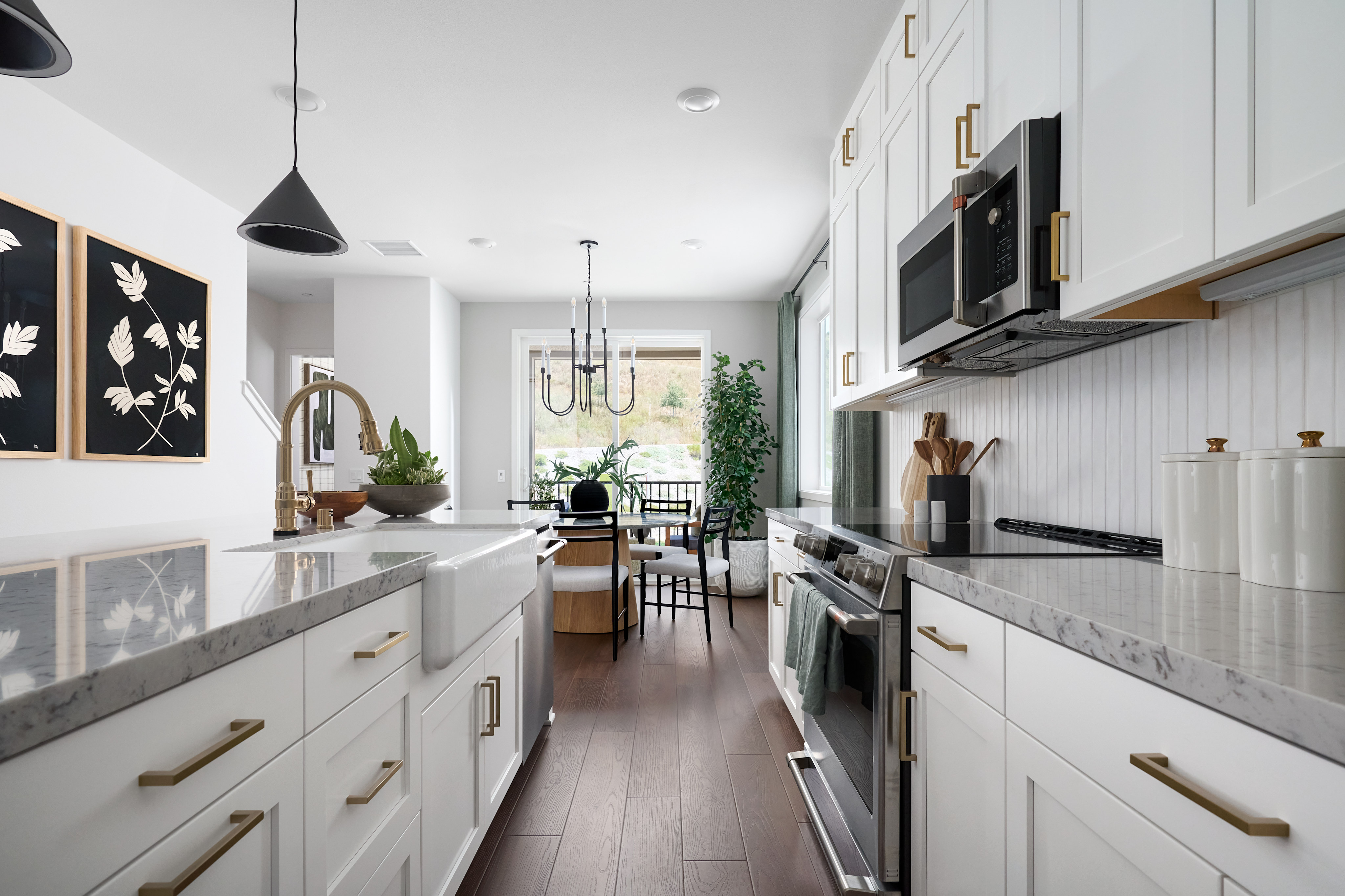 A modern white kitchen with a farmhouse sink in the foreground and a dining area with a chandelier in the background.