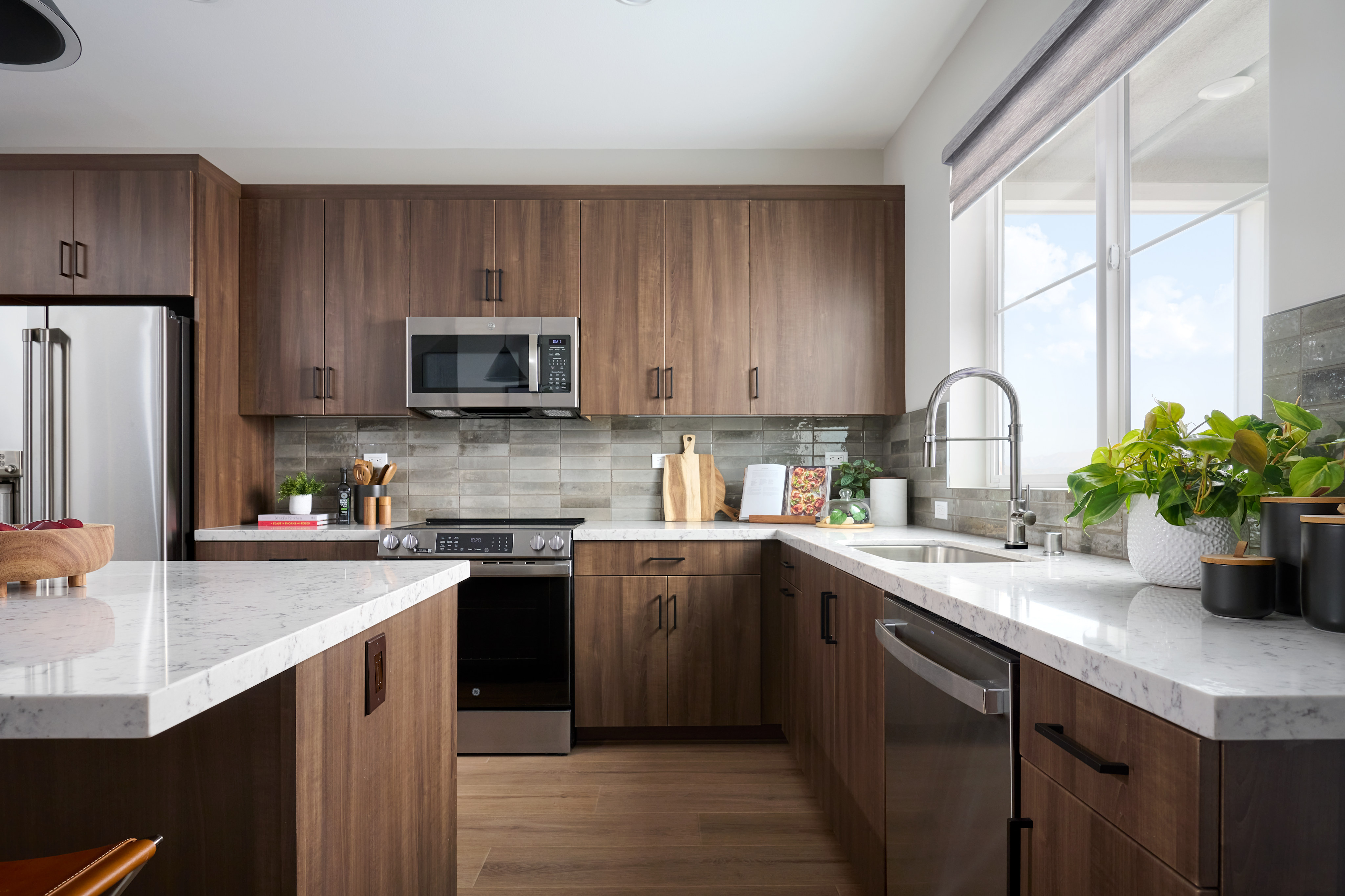 A modern kitchen with a white marble island in the foreground and dark wood cabinets and stainless steel appliances in the background, illuminated by natural light from a large window.