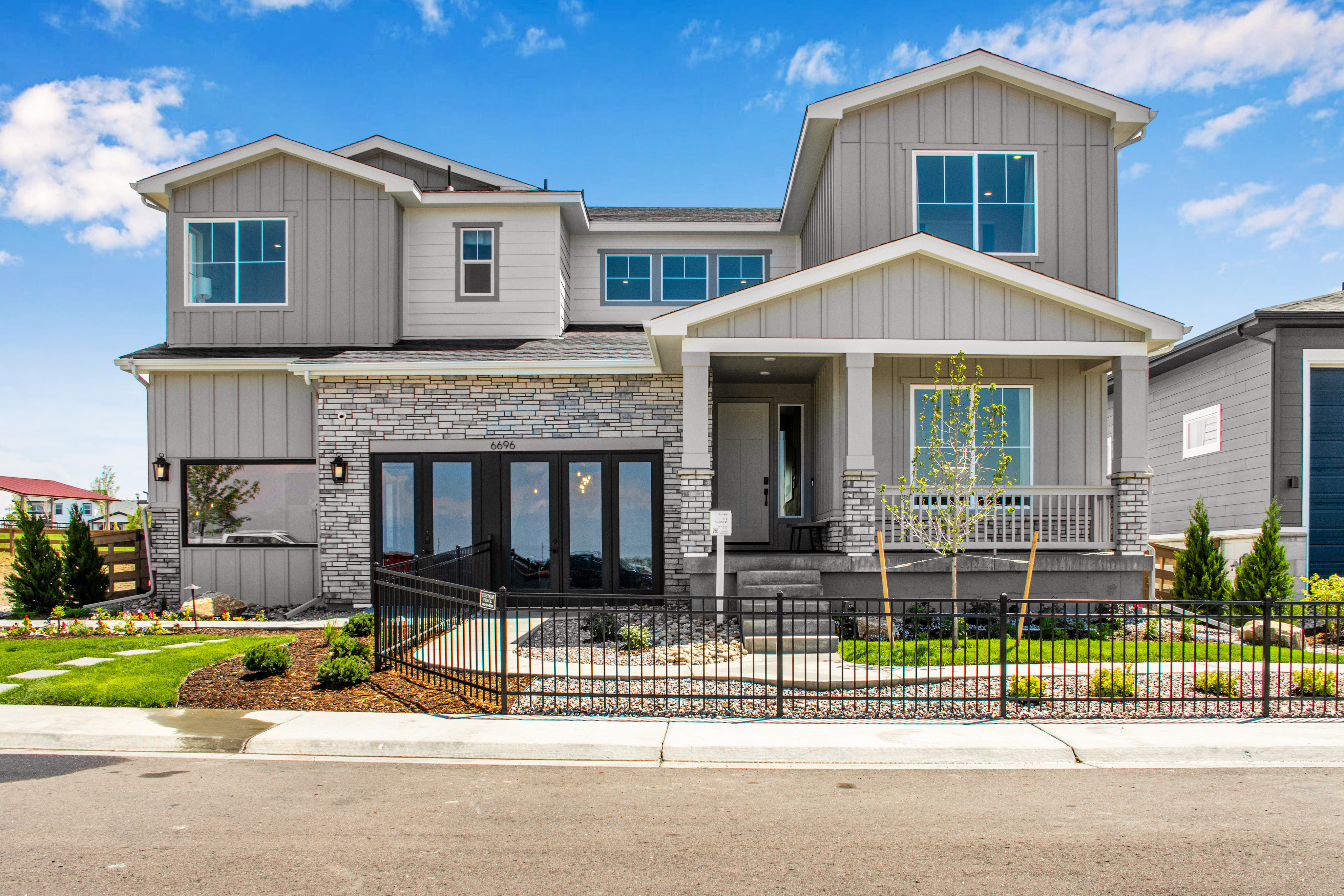 A modern, two-story house with a stone and siding facade stands behind a black metal fence, with a clear blue sky and scattered clouds in the background.