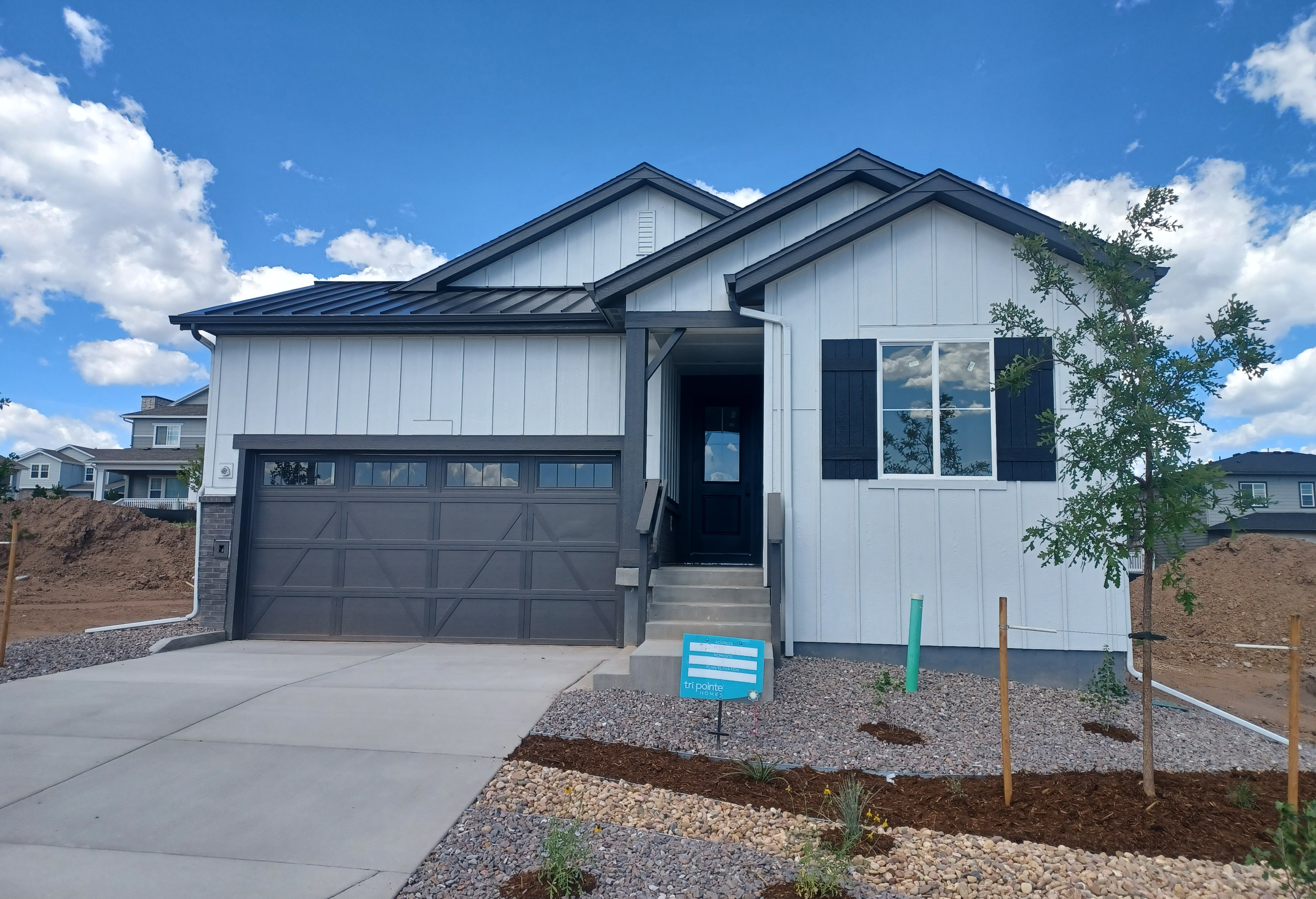 A modern white house with a dark roof and garage door sits on a gravel and mulch landscape in the foreground, with a bright blue sky and scattered clouds in the background.