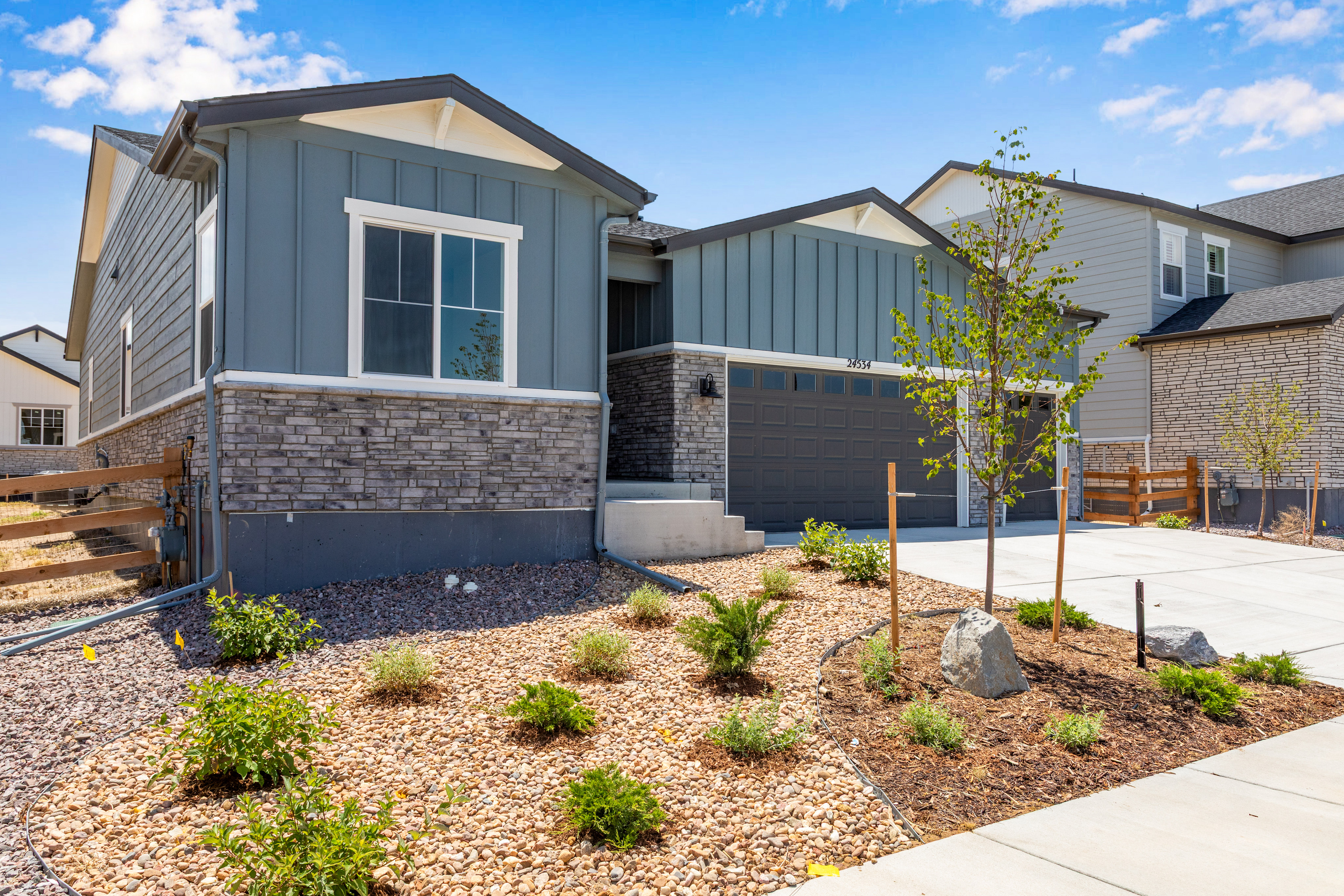 A modern blue and gray house with a xeriscaped front yard sits under a bright blue sky with fluffy clouds.