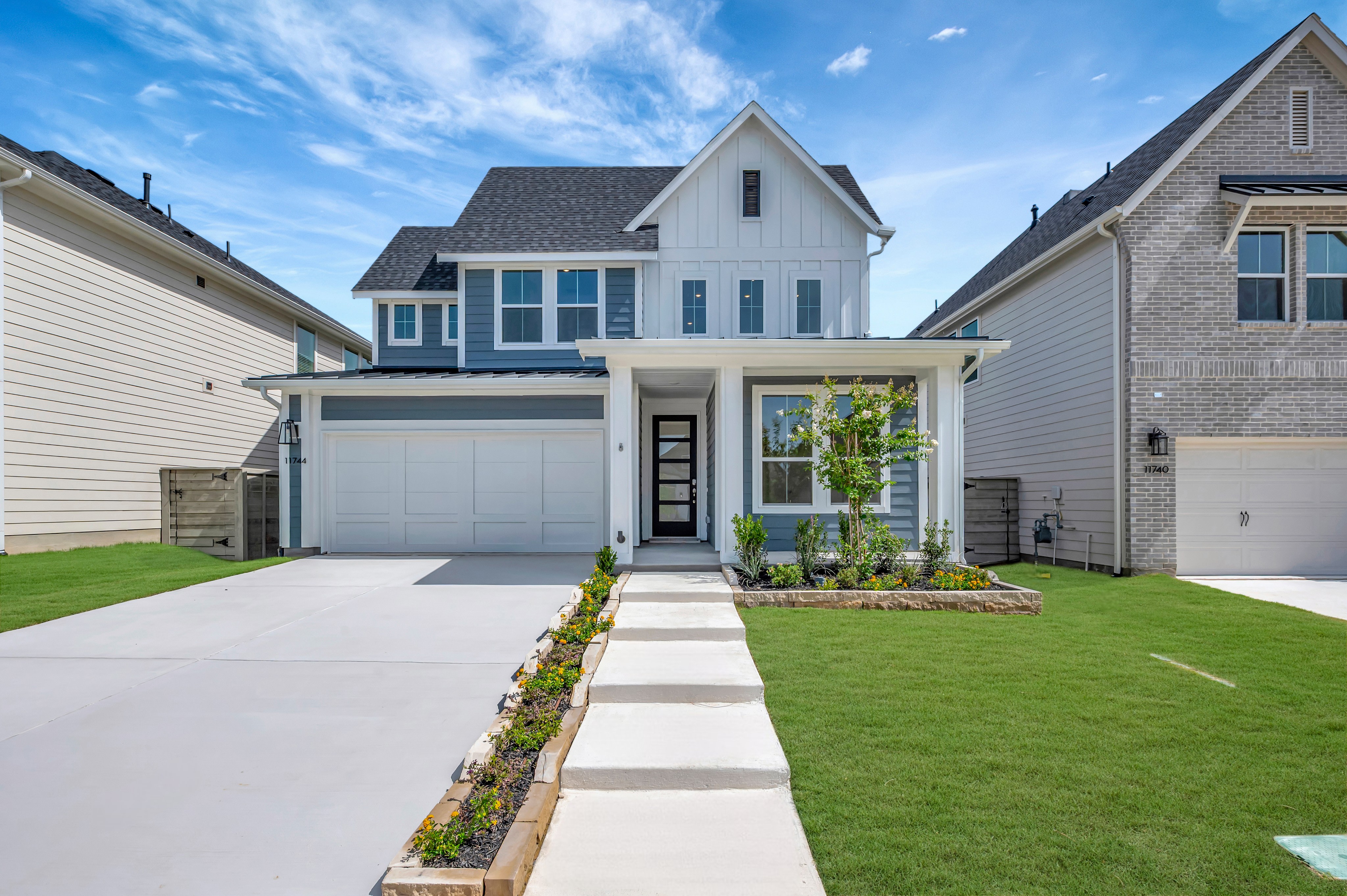 A modern white and blue house with a manicured lawn and walkway in the foreground is surrounded by similar suburban homes under a bright blue sky.