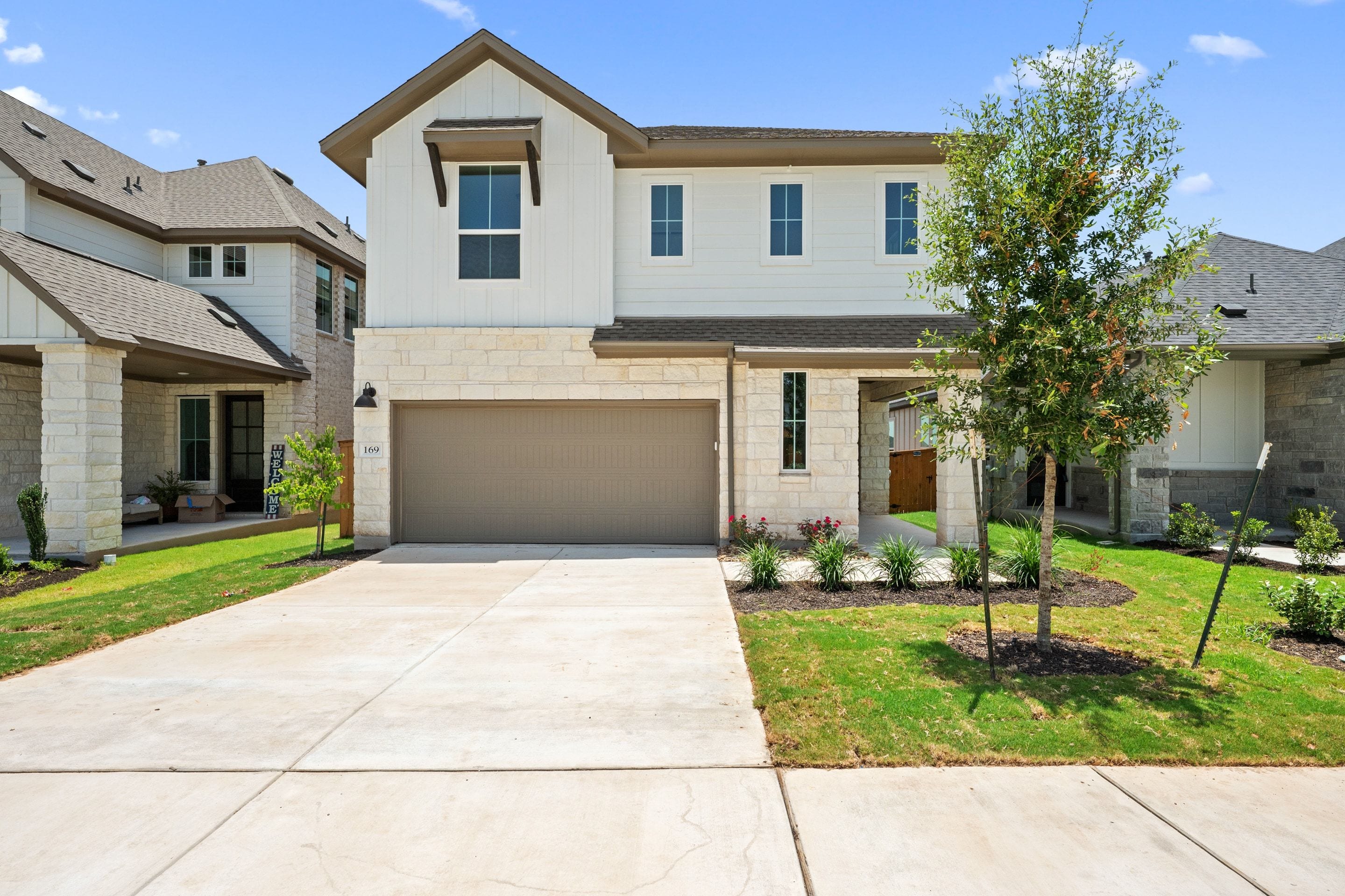 A modern two-story townhouse with a stone and white facade and a two-car garage sits on a manicured lawn with a young tree in the foreground, while other similar townhouses line the street in the background under a clear blue sky.