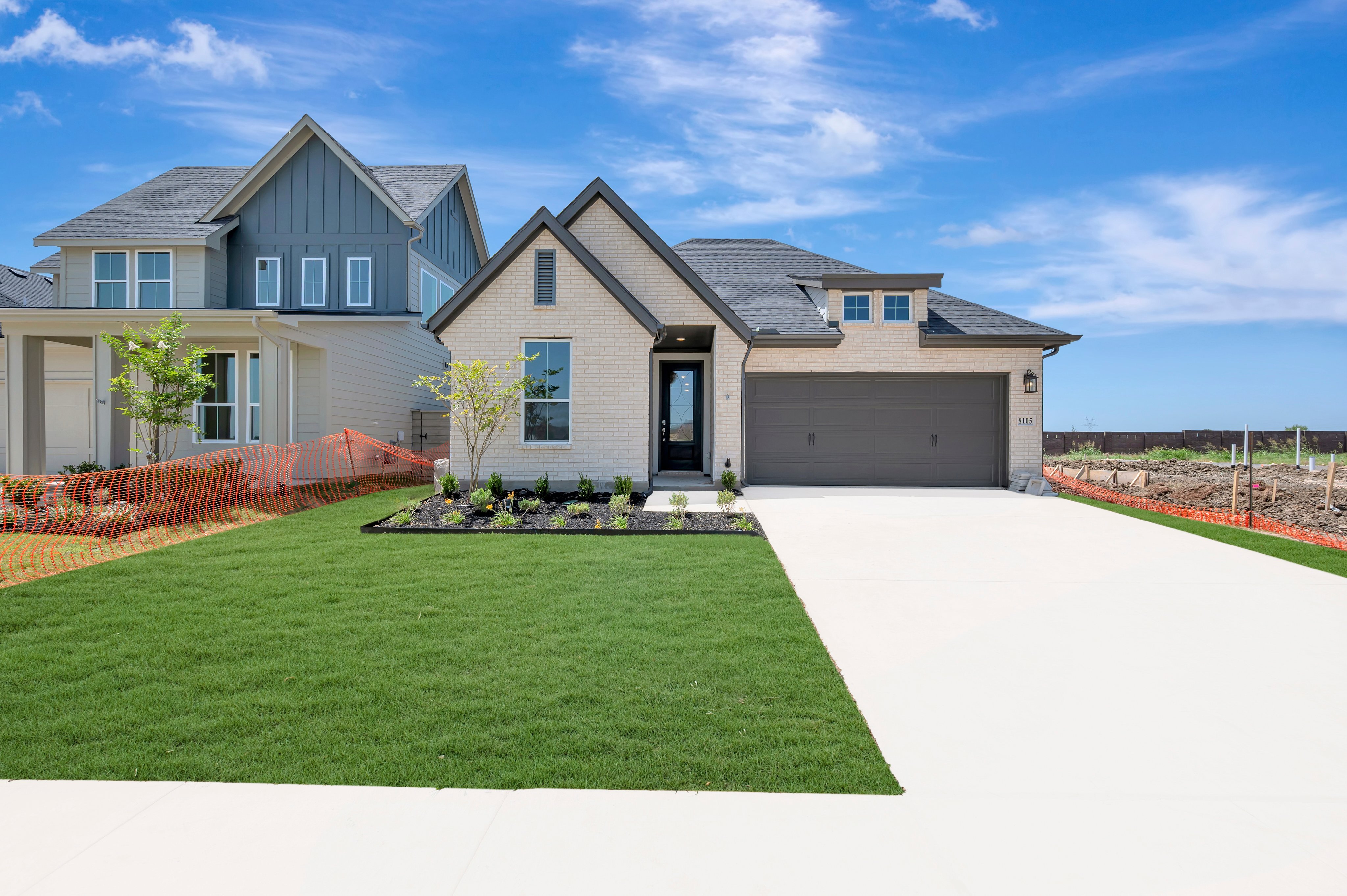 A modern beige brick house with a gray roof and a two-car garage sits on a manicured lawn in the foreground, while a blue sky with scattered clouds forms the background.
