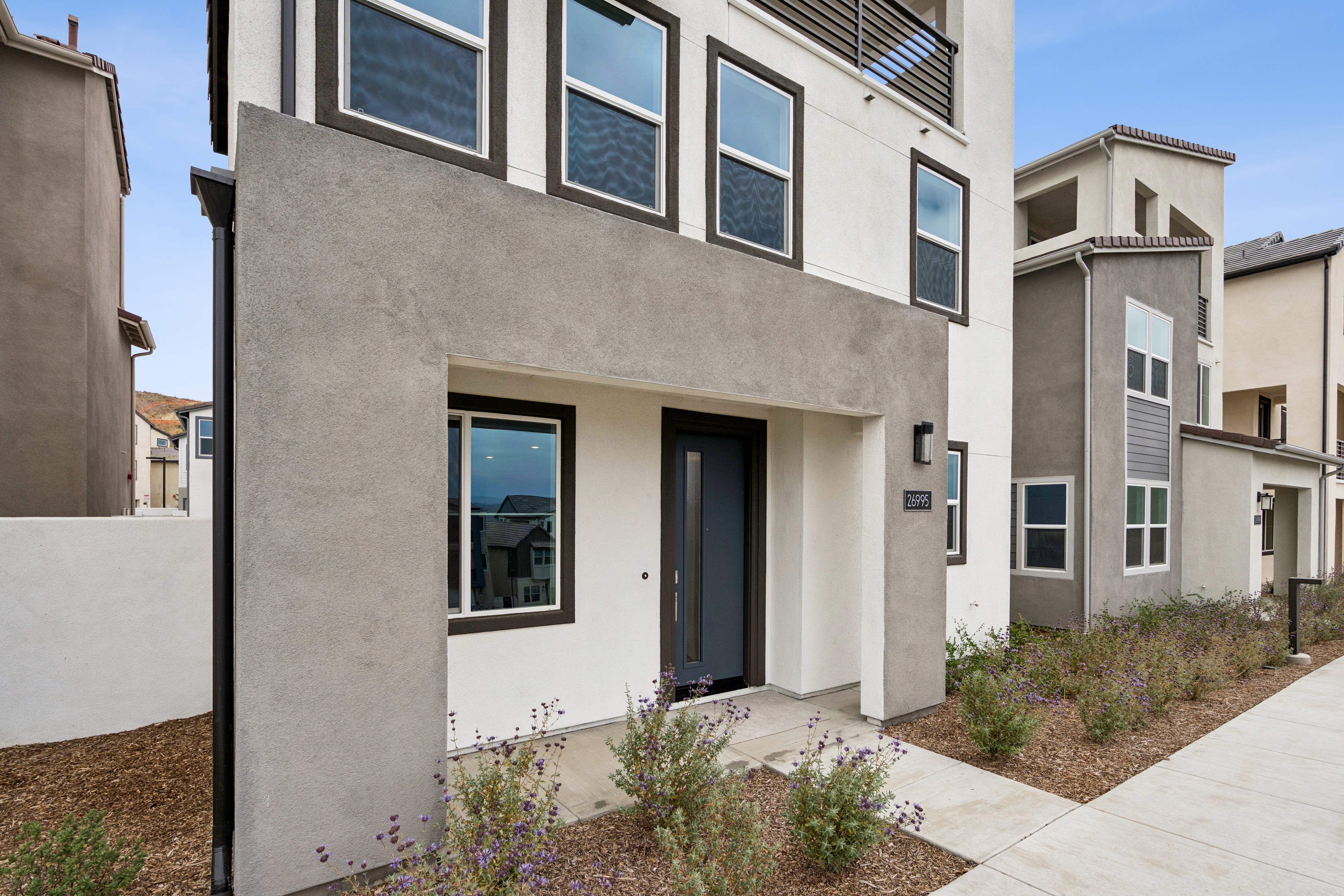 A modern townhouse with a gray stucco entryway and a paved walkway is flanked by landscaping and other similar homes under a clear sky.