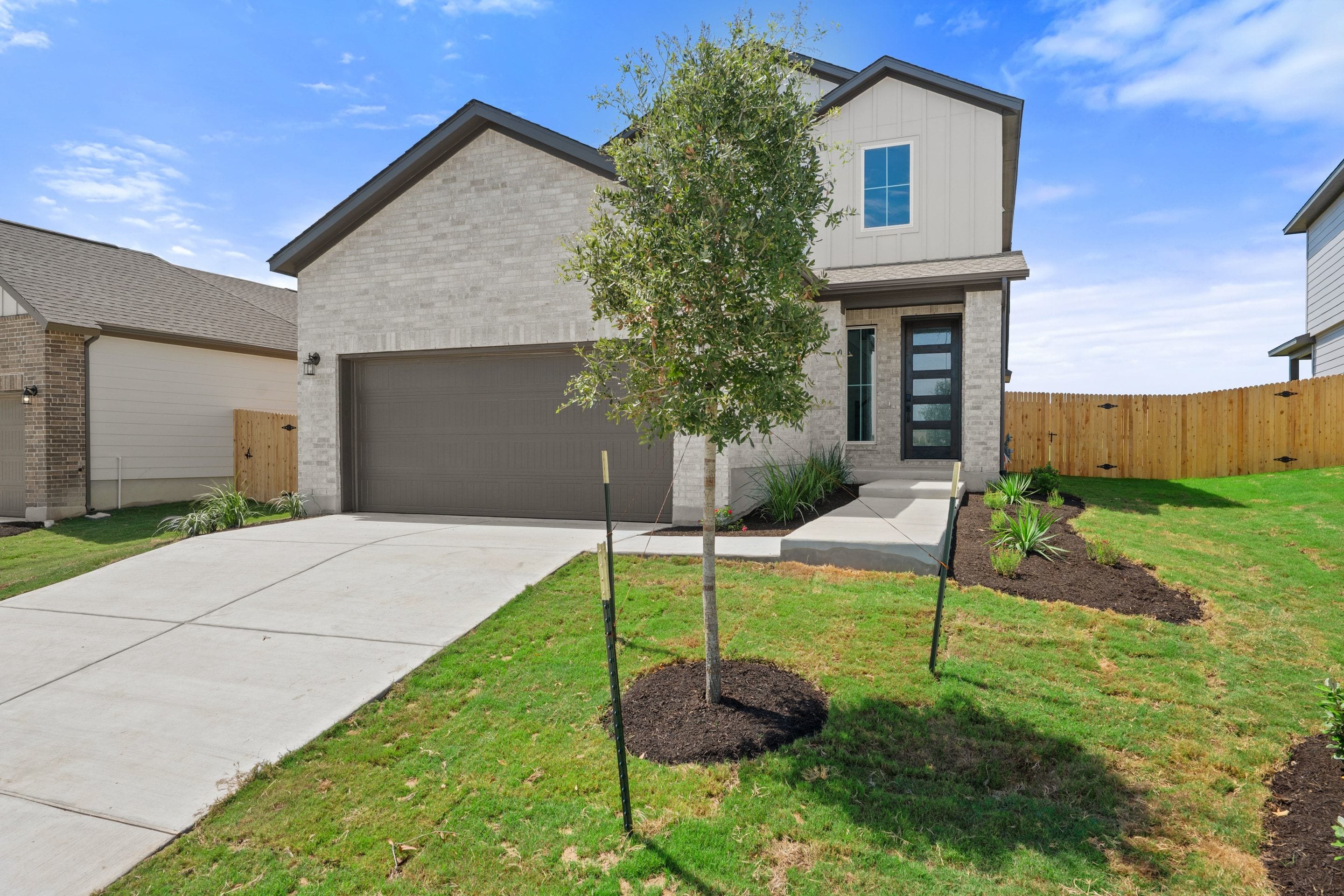 A modern two-story house with a light brick exterior and a dark garage door sits on a manicured lawn with a young tree in the foreground, while a wooden fence and other houses form the background under a blue sky.