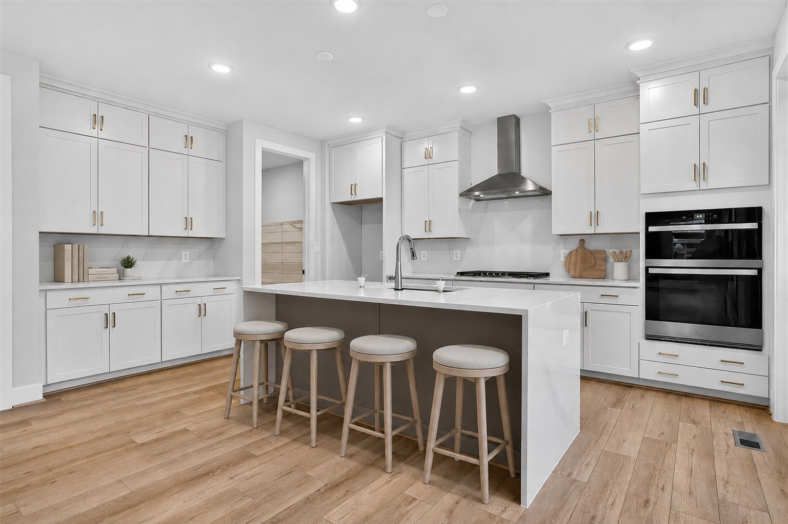A modern white kitchen with a large island and four stools in the foreground, and white cabinetry, stainless steel appliances, and a doorway leading to another room in the background.