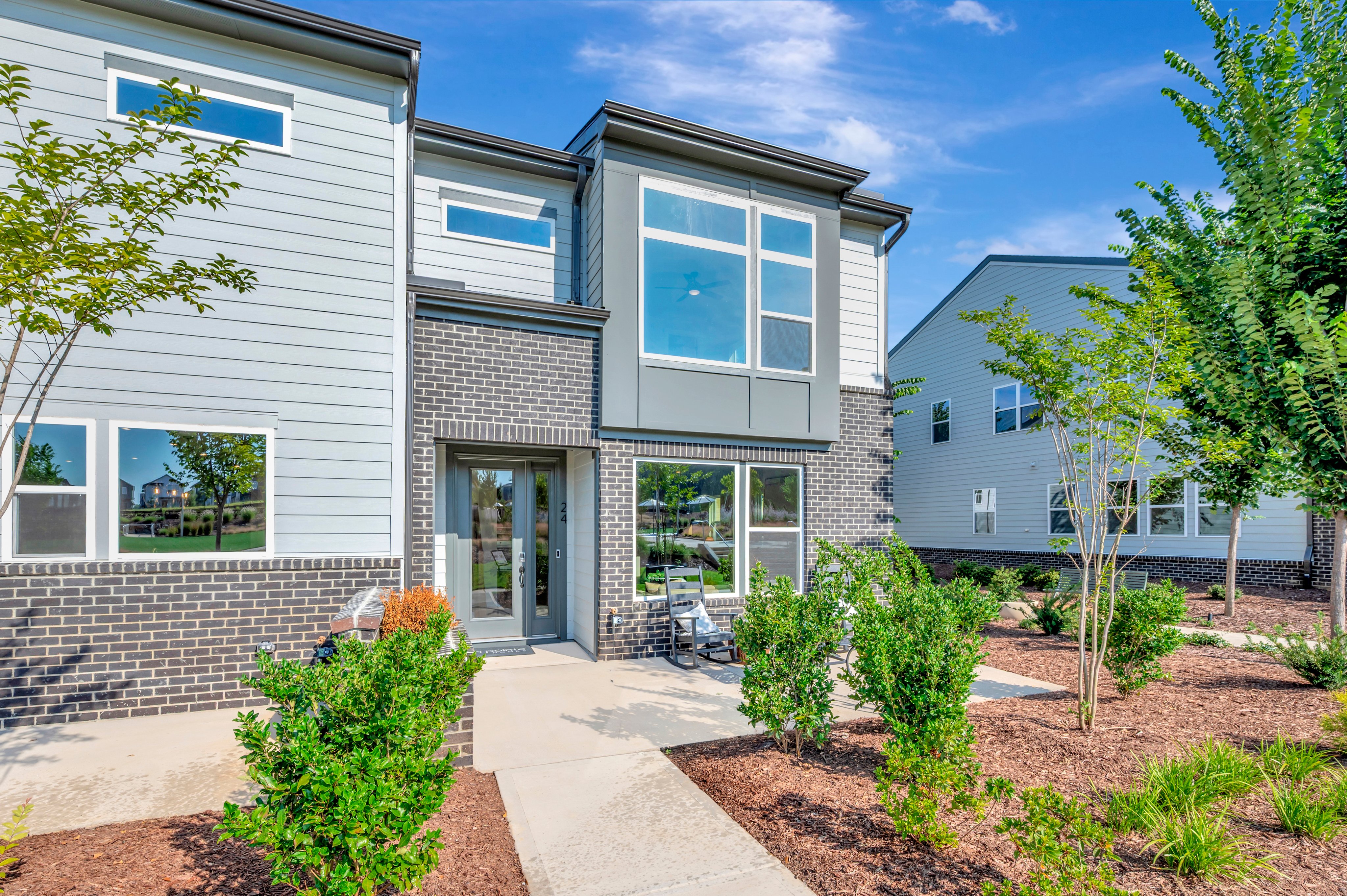 A modern townhouse with a gray brick and siding exterior stands on a paved walkway surrounded by lush green bushes, with other similar houses visible in the background under a bright blue sky.