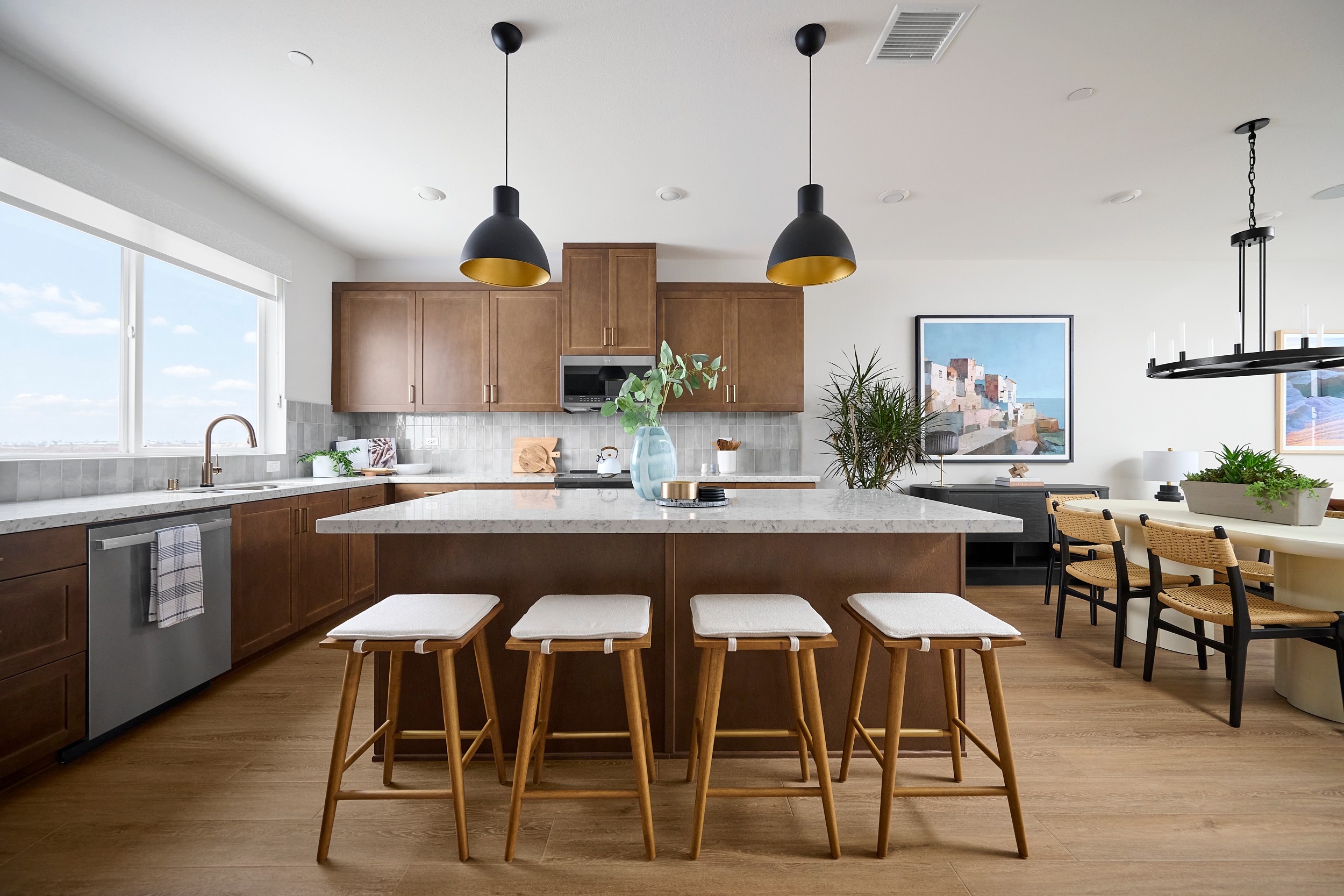 Four white-cushioned stools with wooden legs surround a granite-topped island in the foreground, while a modern kitchen with wooden cabinets and a large window is visible in the background.