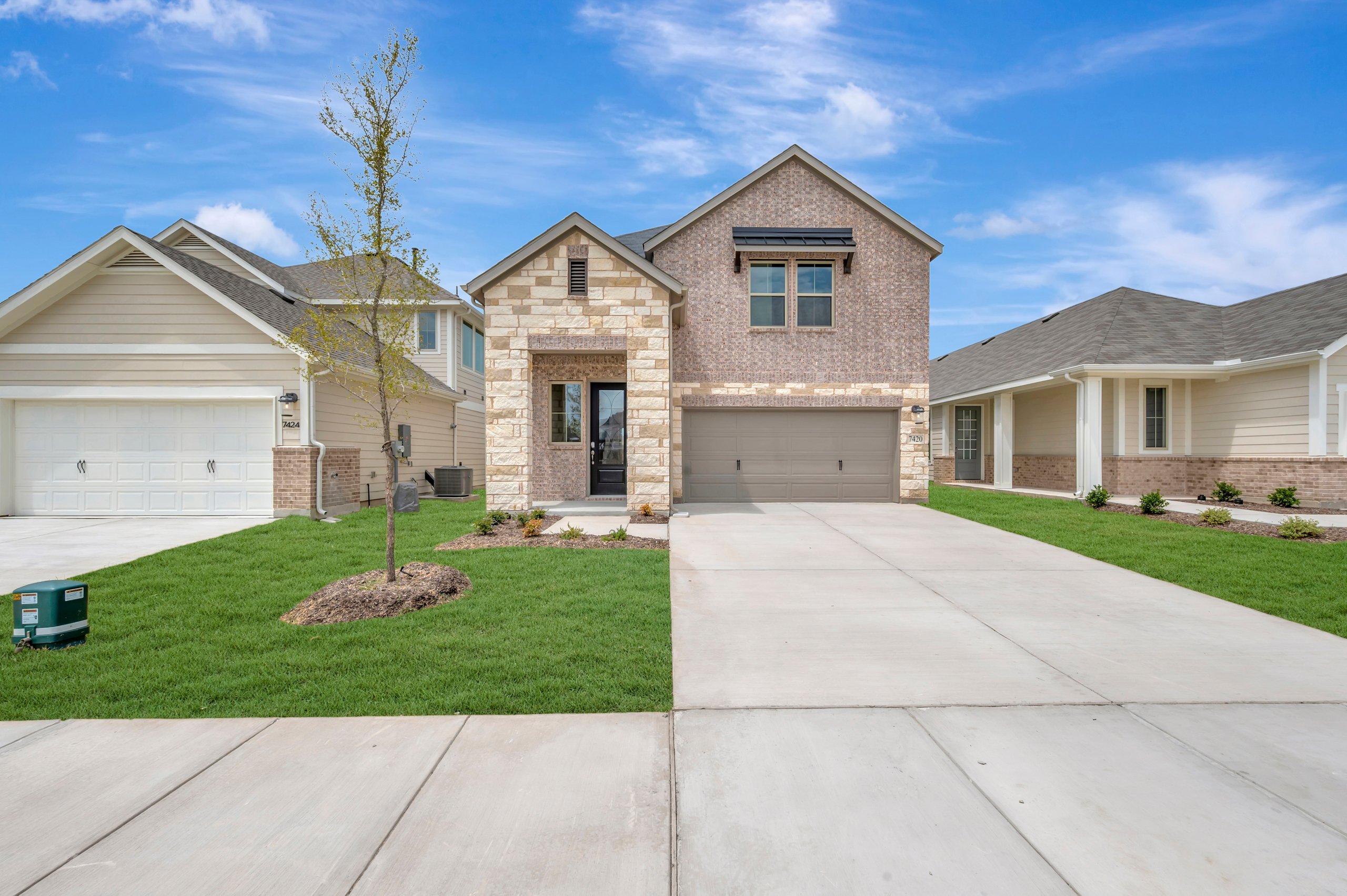 A modern brick and stone house with a manicured lawn and driveway sits under a bright blue sky with scattered clouds, flanked by similar suburban homes.