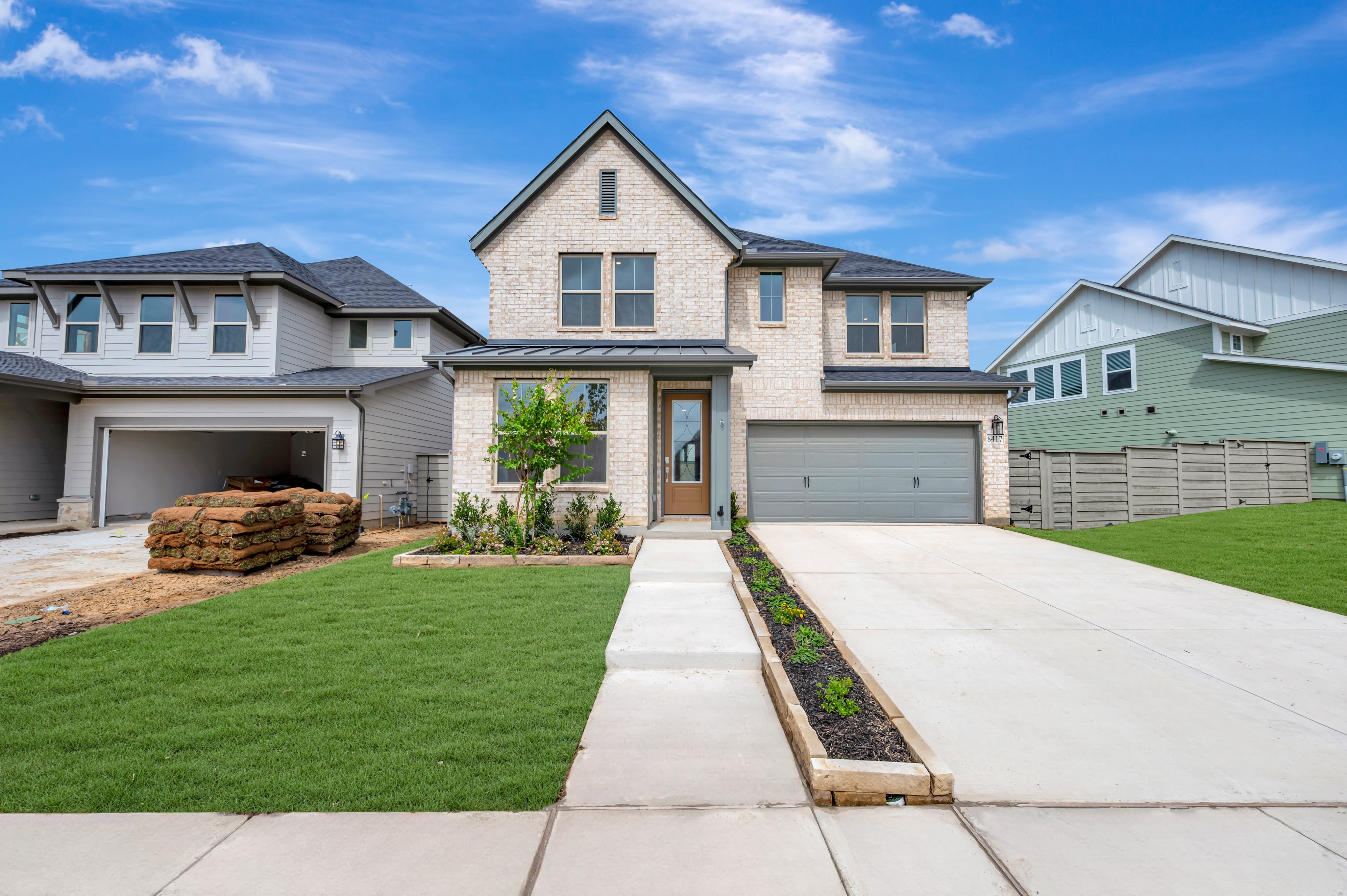 A modern brick house with a manicured lawn and driveway sits under a bright blue sky with fluffy clouds, flanked by other contemporary homes.