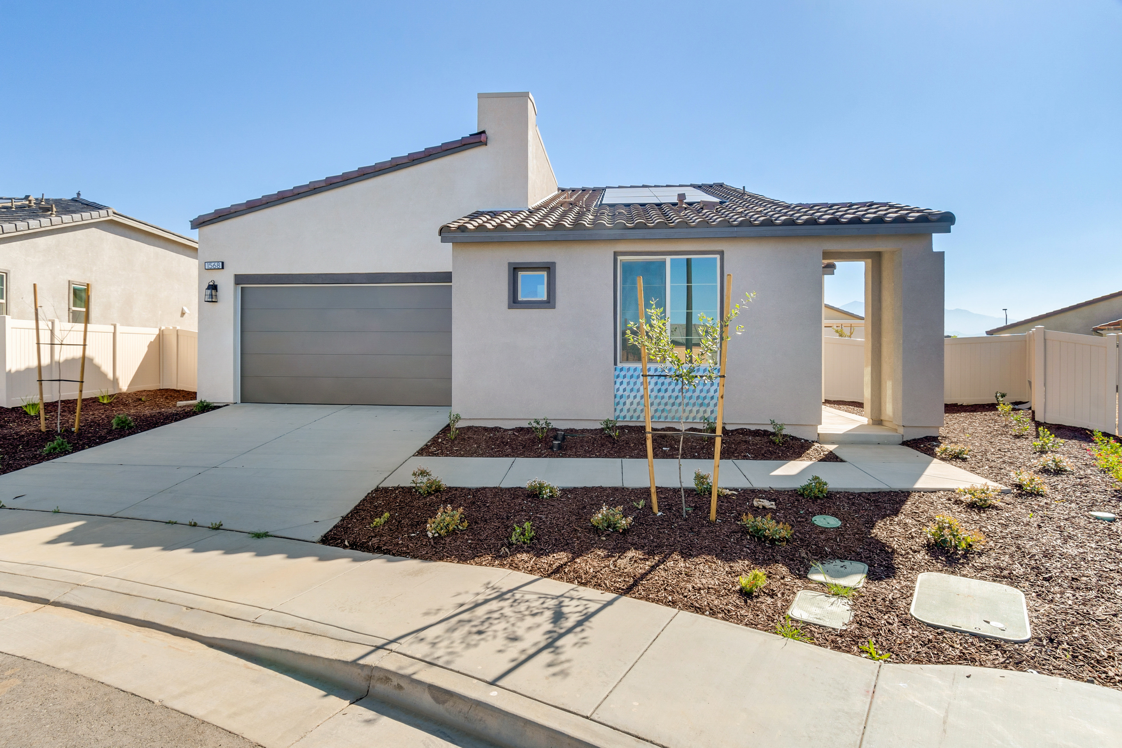 A modern stucco house with a gray garage door and a tiled roof sits behind a landscaped yard with young trees, while other houses and a clear blue sky form the background.