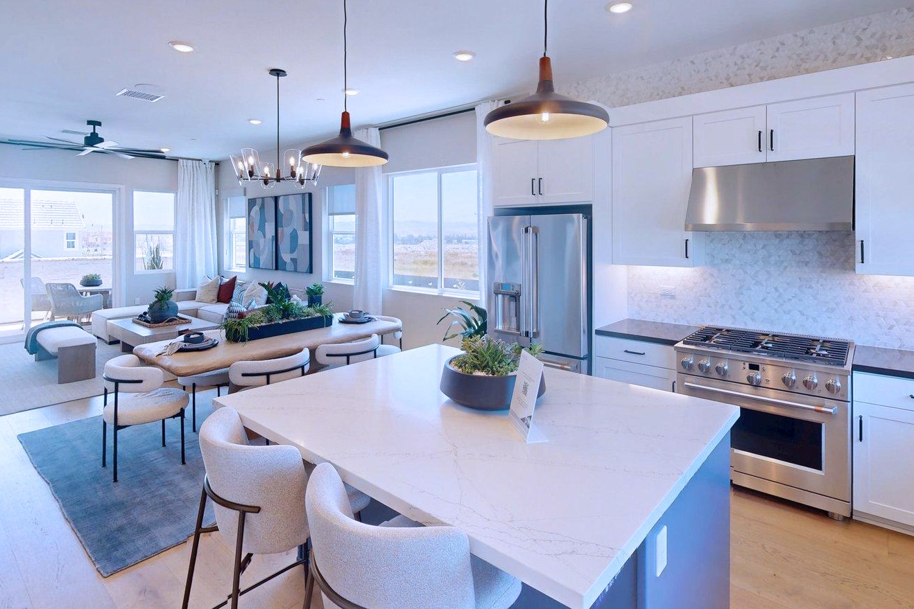 A modern kitchen island with bar stools in the foreground leads to a spacious living area with a couch and dining table, all bathed in natural light from large windows overlooking a distant landscape.