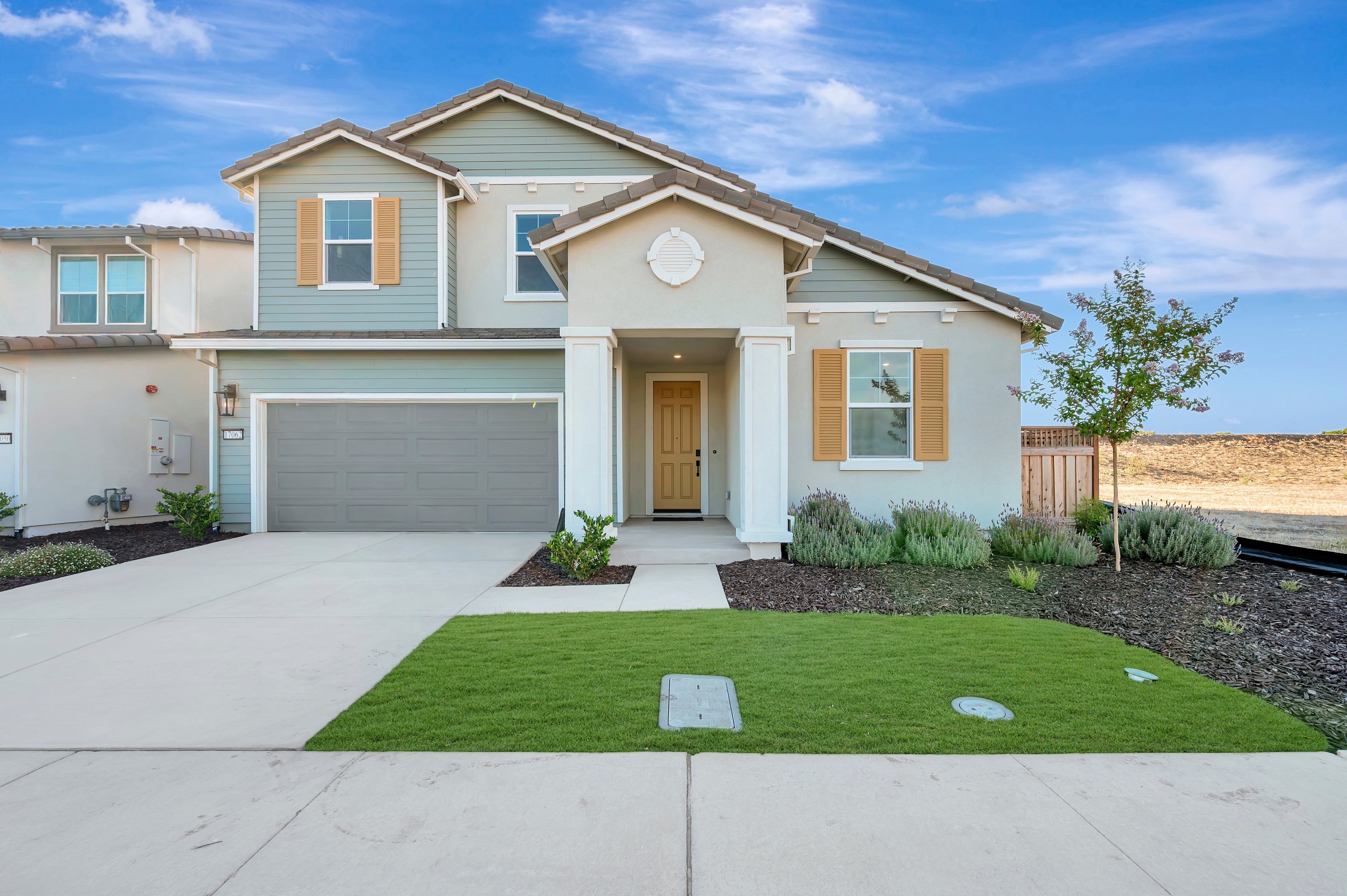 A modern two-story house with a well-manicured lawn in the foreground sits under a bright blue sky with scattered clouds, while a dry, undeveloped landscape is visible in the background.