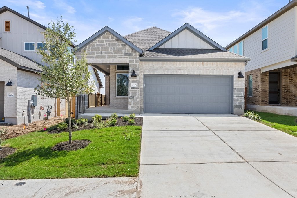 A modern house with a gray garage door and stone accents sits on a manicured lawn, with neighboring houses visible in the background under a cloudy sky.