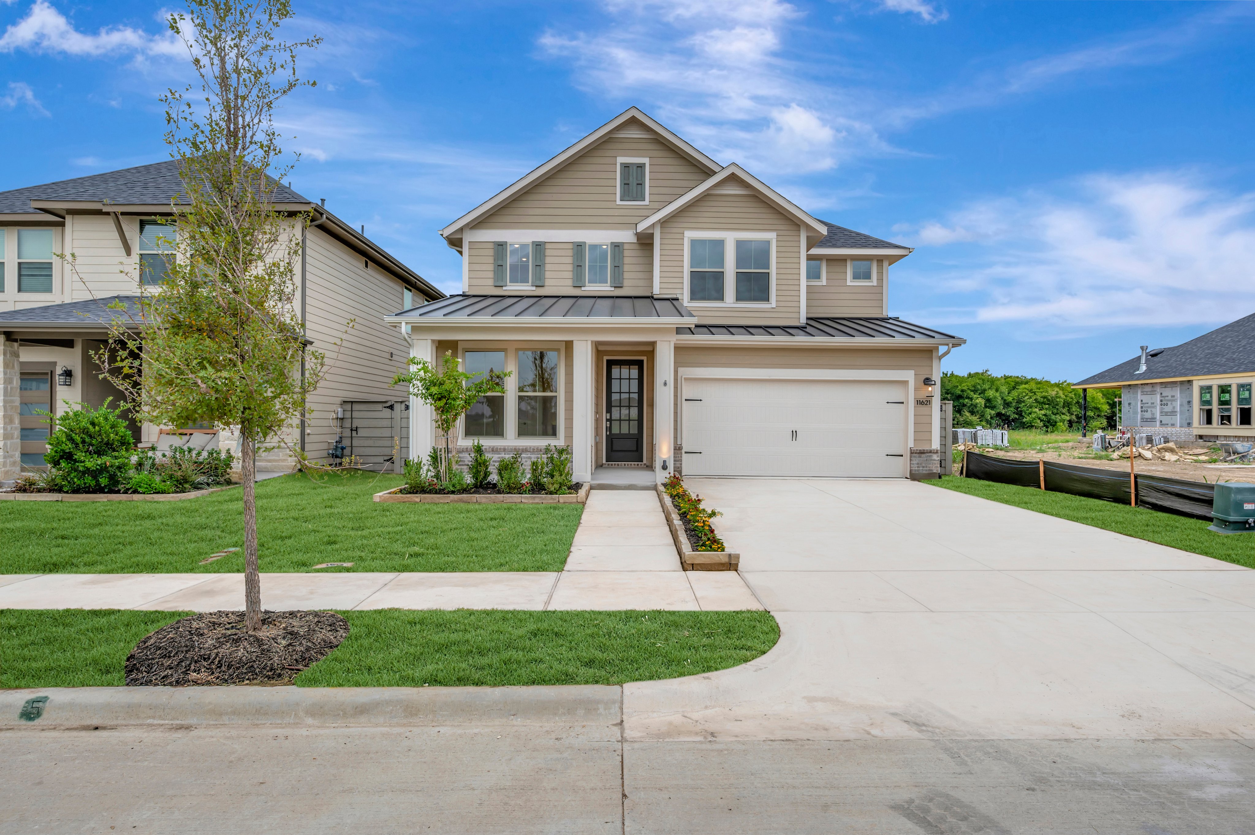 A tan two-story house with a white garage door and a paved driveway sits on a manicured lawn, with a bright blue sky and distant construction in the background.