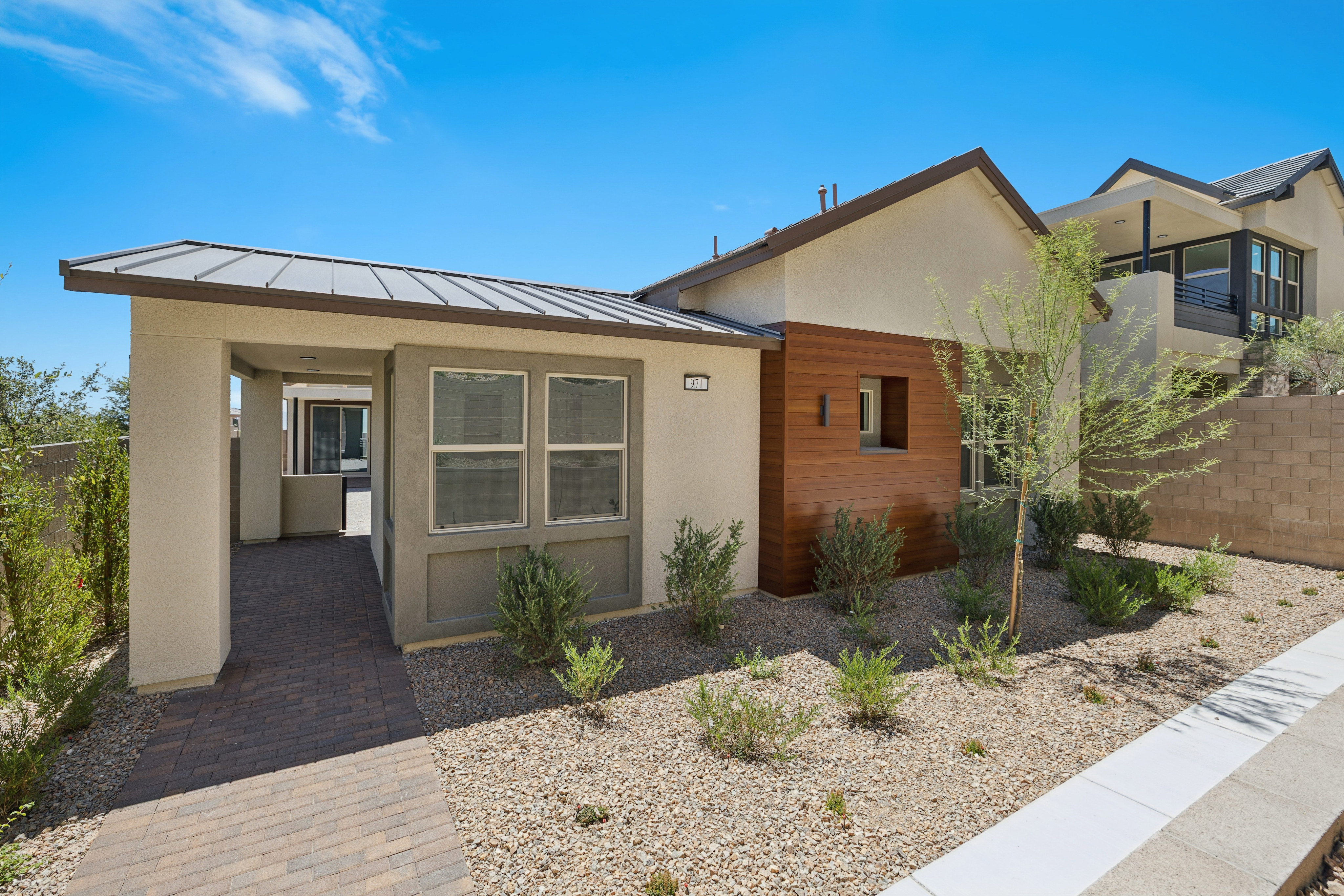 A modern house with a light-colored facade and wood accents sits in the foreground, surrounded by drought-tolerant landscaping, while other houses are visible on a hillside in the background under a clear blue sky.