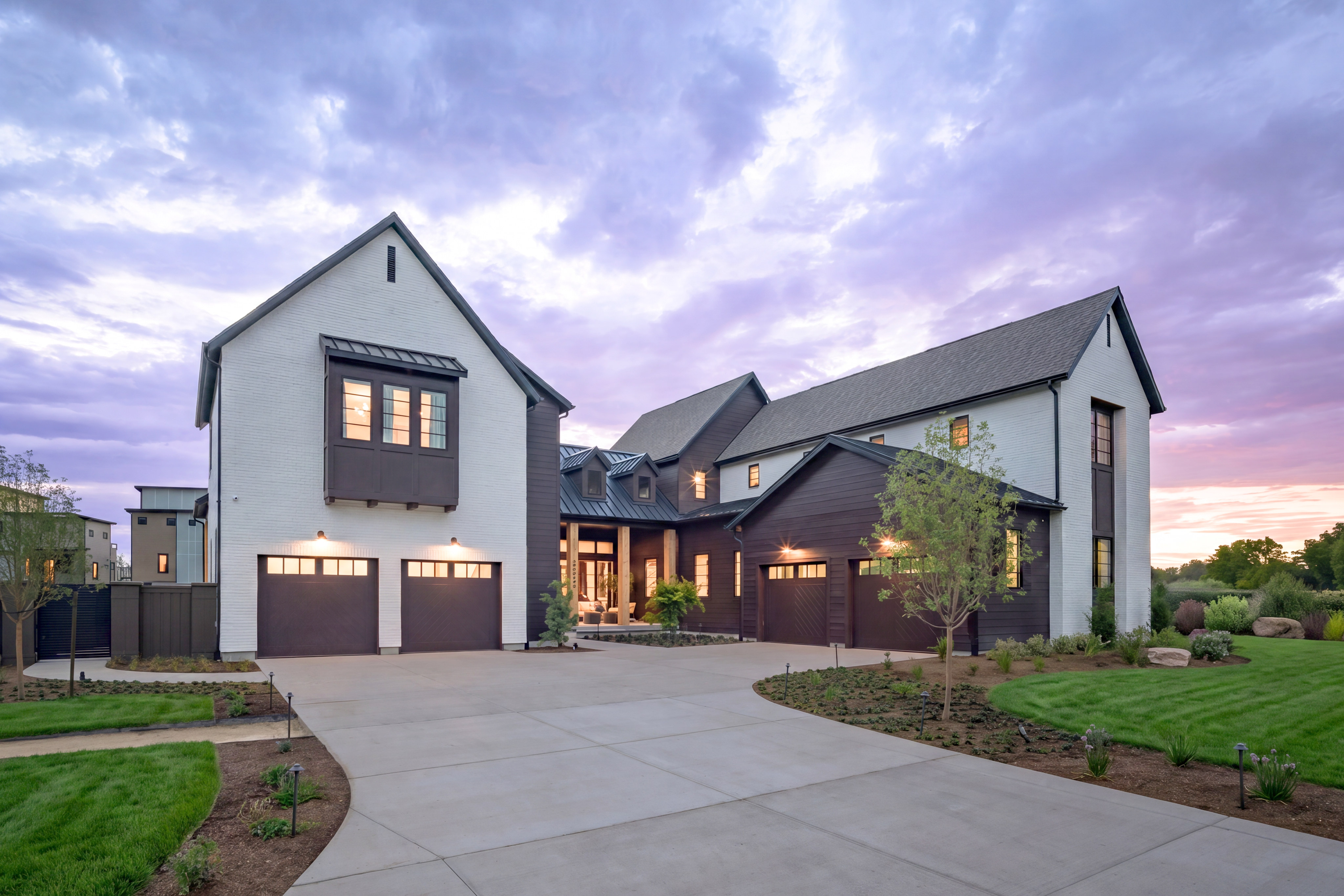 A modern white and dark wood house with a wide driveway in the foreground sits under a dramatic purple and pink sky.
