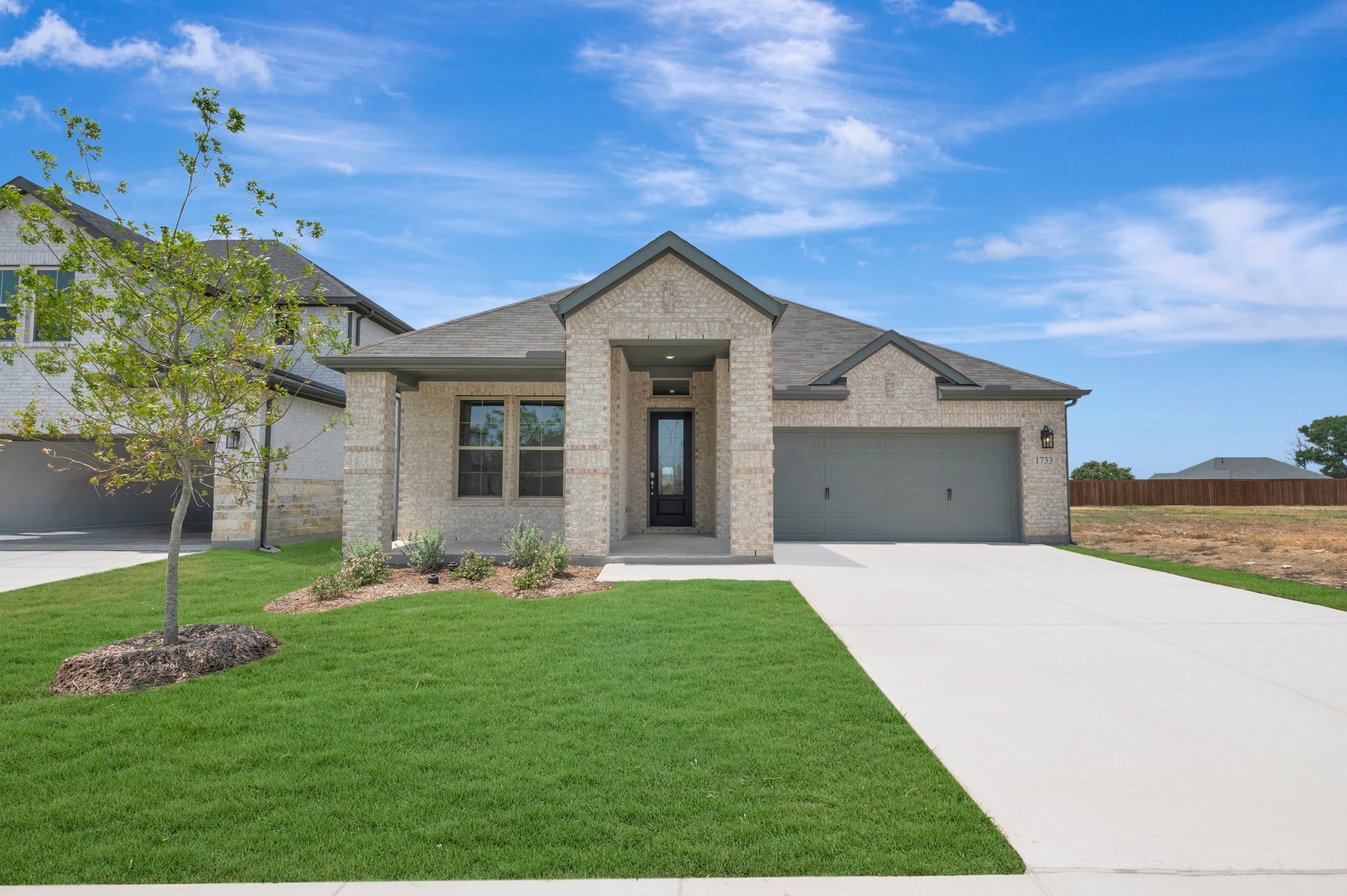 A modern brick house with a manicured lawn in the foreground sits under a bright blue sky with scattered clouds, while a neighboring house and undeveloped land are visible in the background.