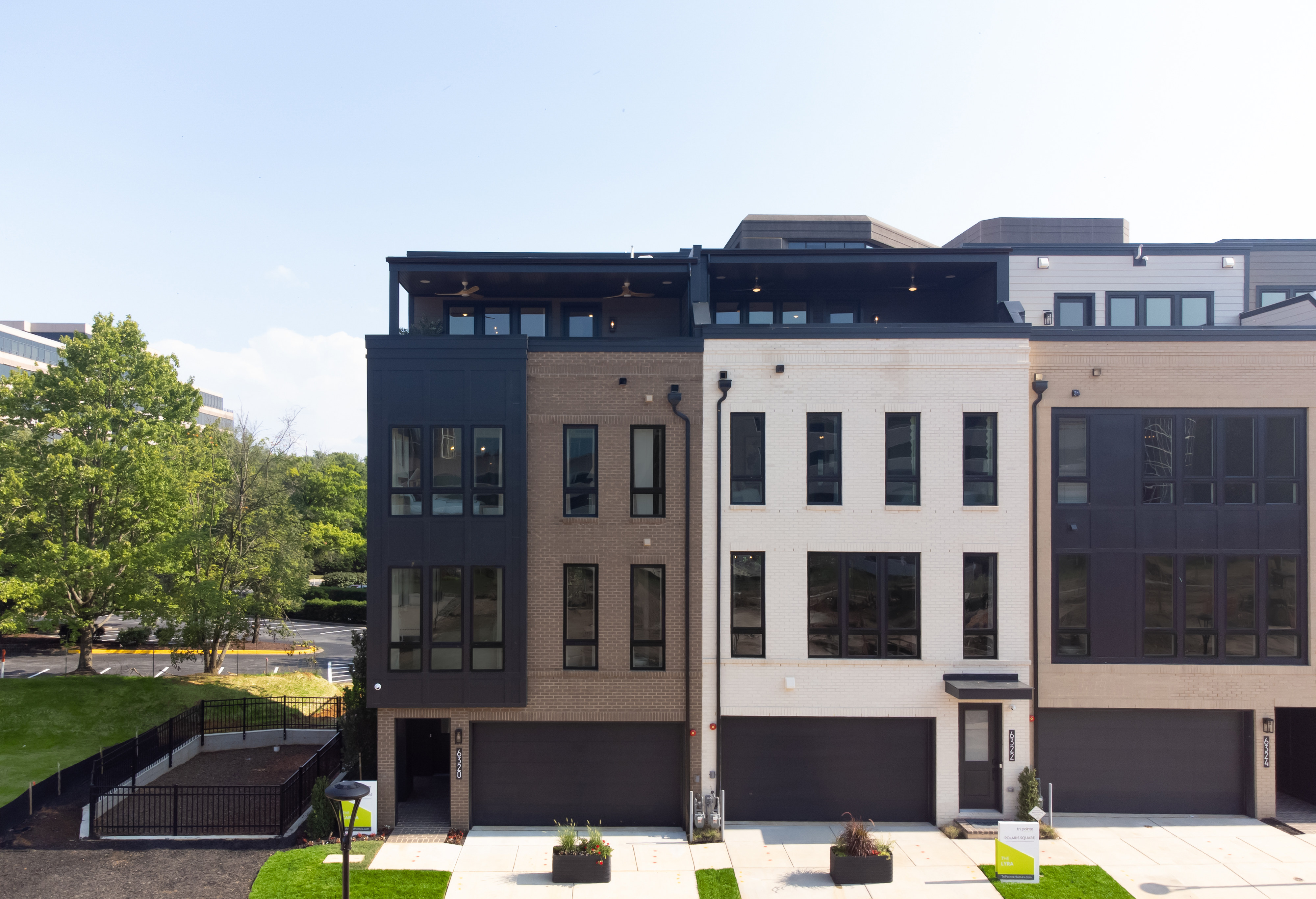 Modern townhouses with dark garages and large windows stand in the foreground, while a lush green lawn and trees are visible in the background under a clear sky.