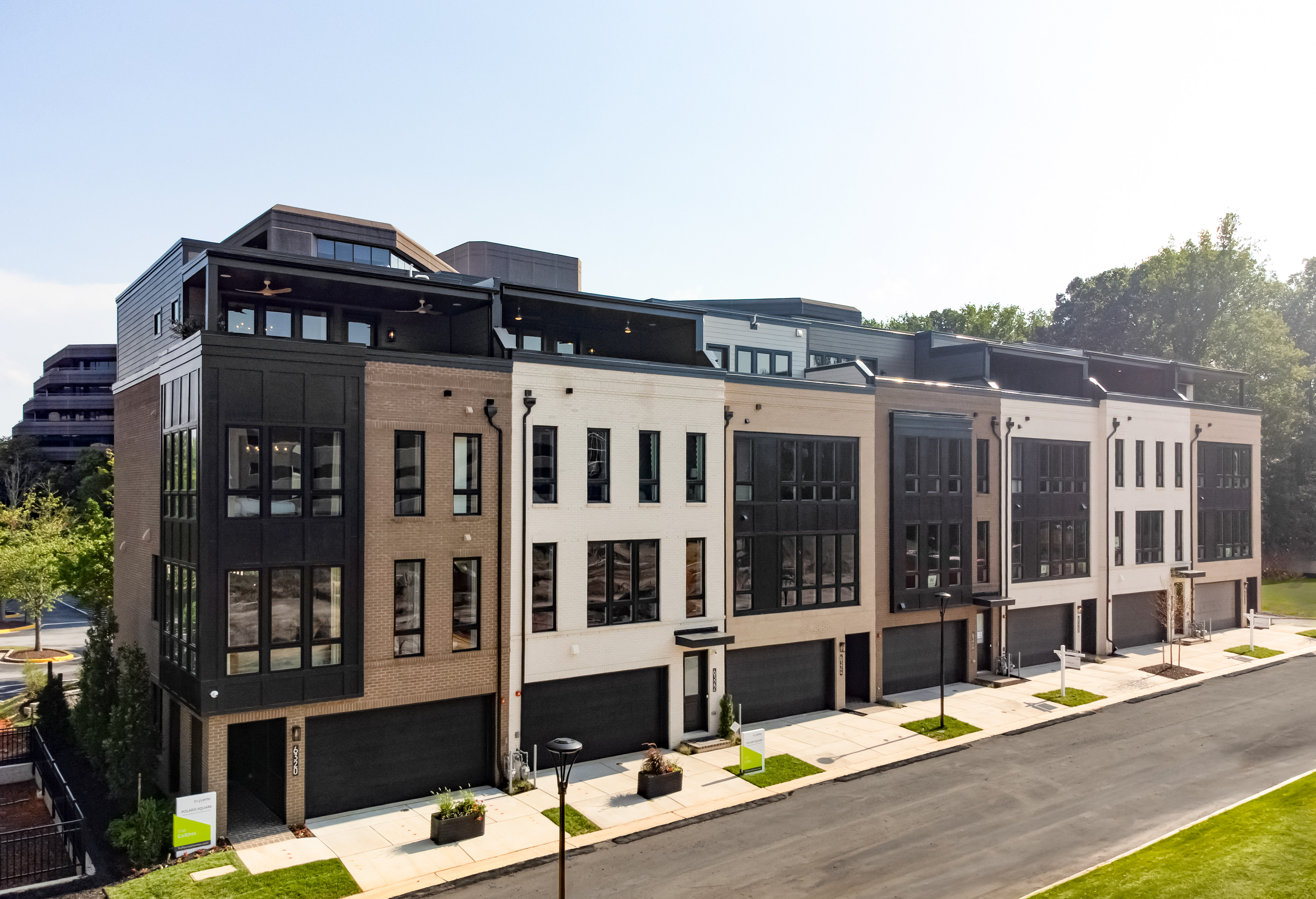 A row of modern townhouses with large windows and garages lines a street, with trees and a distant building in the background.