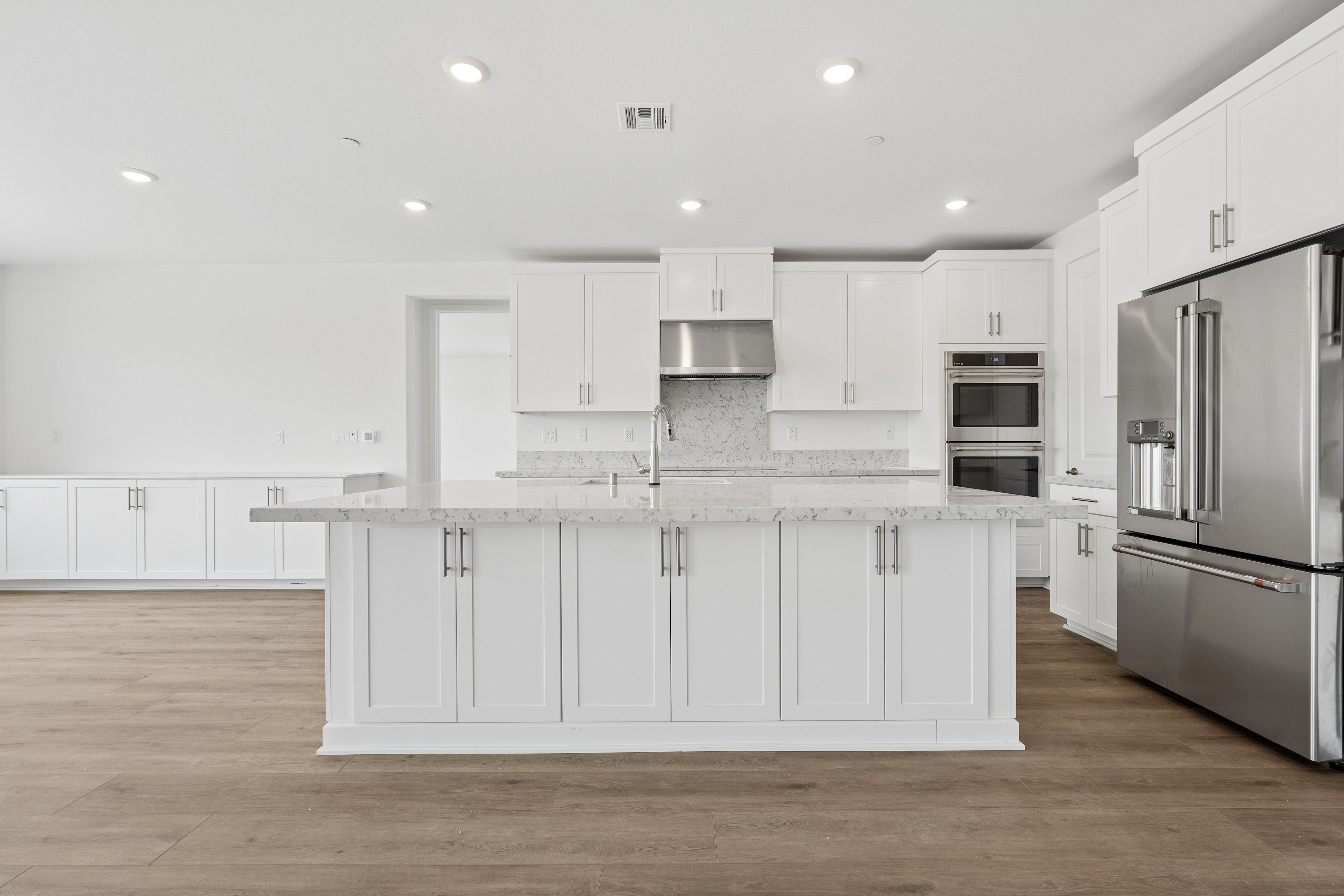 A modern white kitchen with a large island in the foreground and a stainless steel refrigerator and built-in ovens in the background.