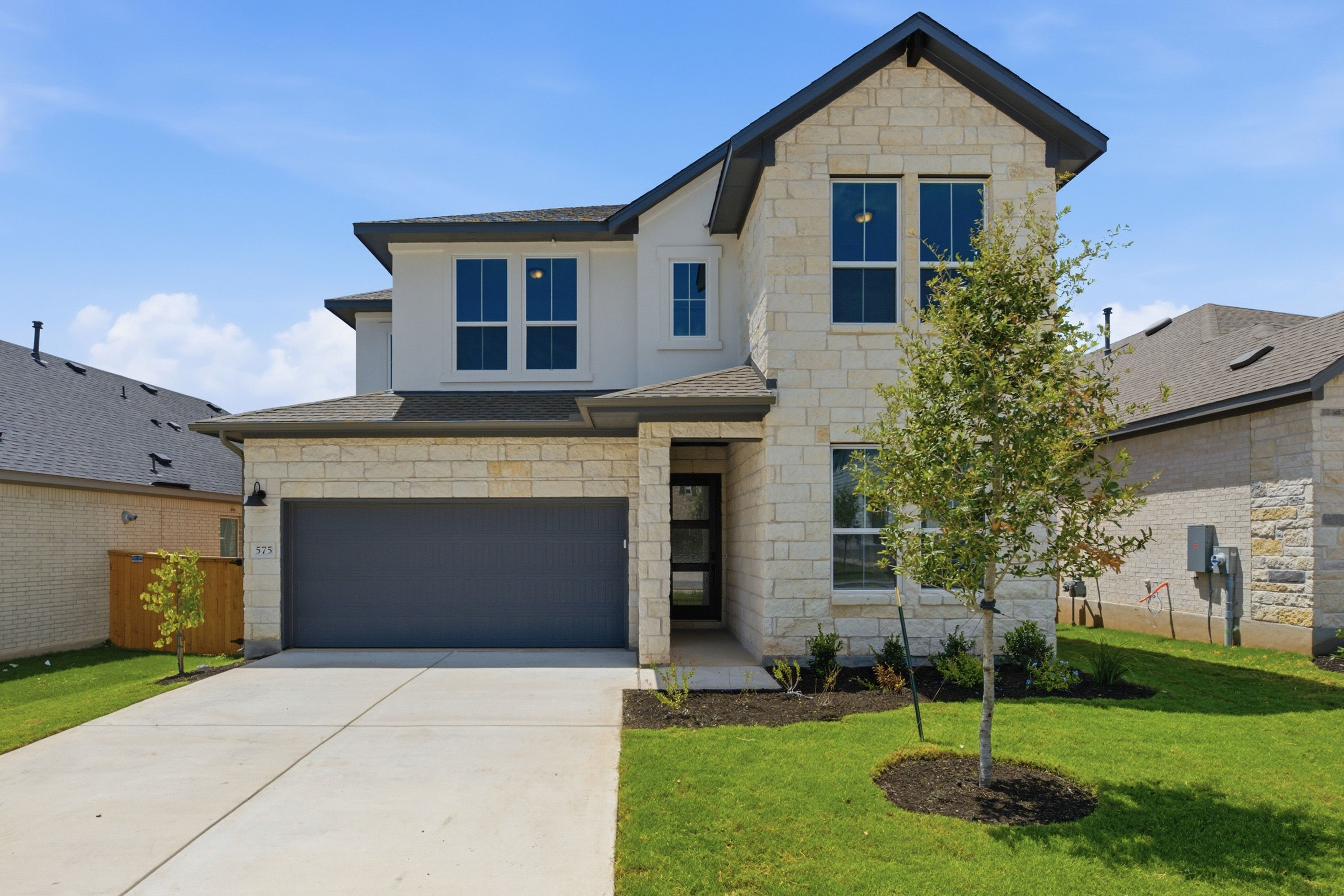 A modern two-story house with a stone facade and a gray garage door sits on a manicured lawn, with neighboring houses and a clear blue sky in the background.