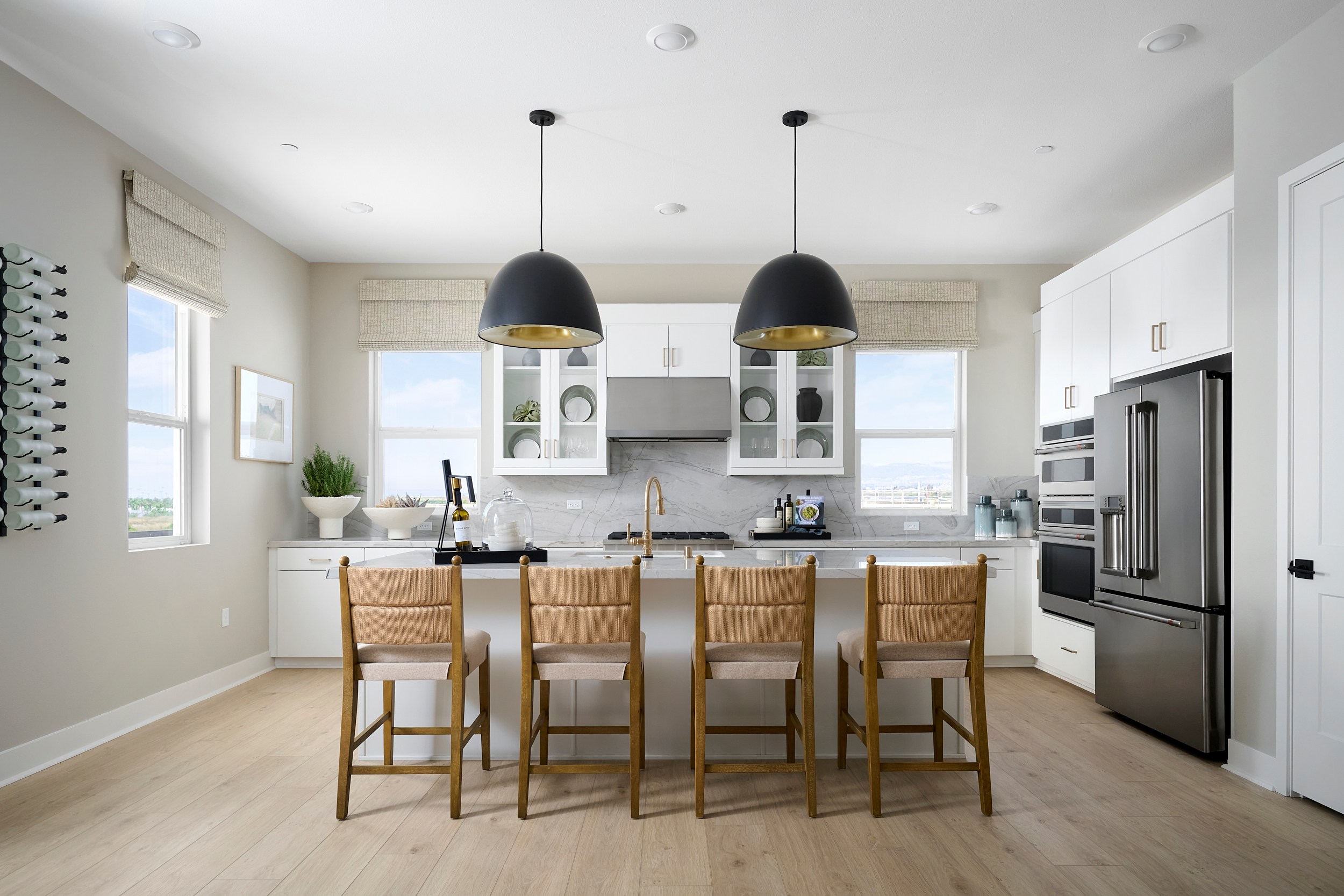 Four woven-seat bar stools are arranged at a white island with a marble countertop, while a stainless steel refrigerator and white cabinets fill the bright, modern kitchen background.
