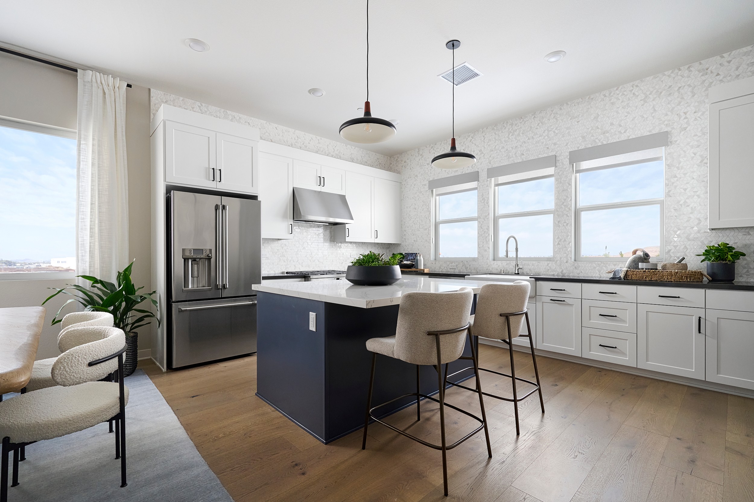 A modern kitchen with a dark blue island and bar stools in the foreground, and white cabinets and windows with a bright view in the background.
