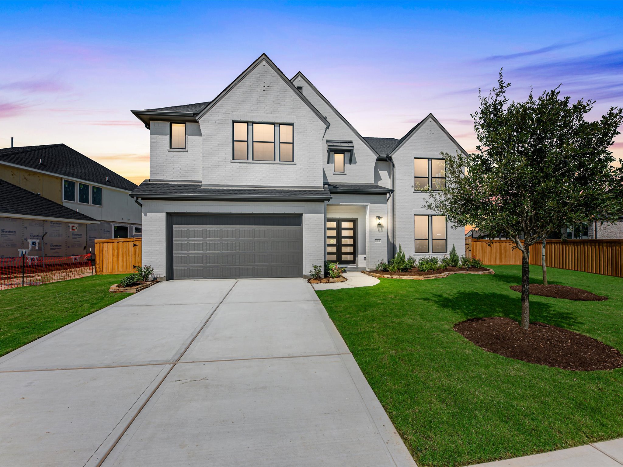A modern white brick house with a gray garage door sits on a manicured lawn in front of a twilight sky with neighboring houses visible in the distance.