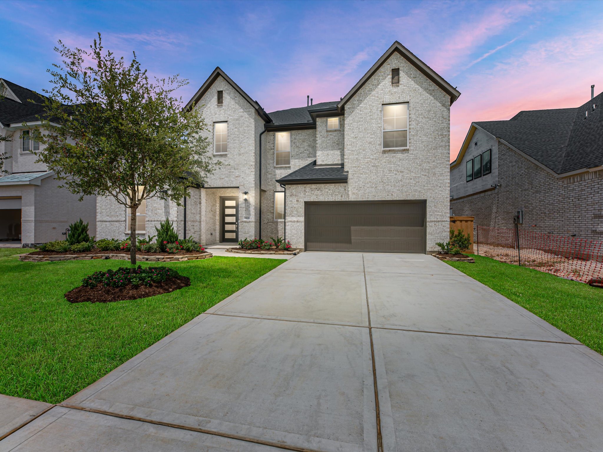 A modern white brick house with a large driveway sits in the foreground, while neighboring houses and a colorful twilight sky form the background.