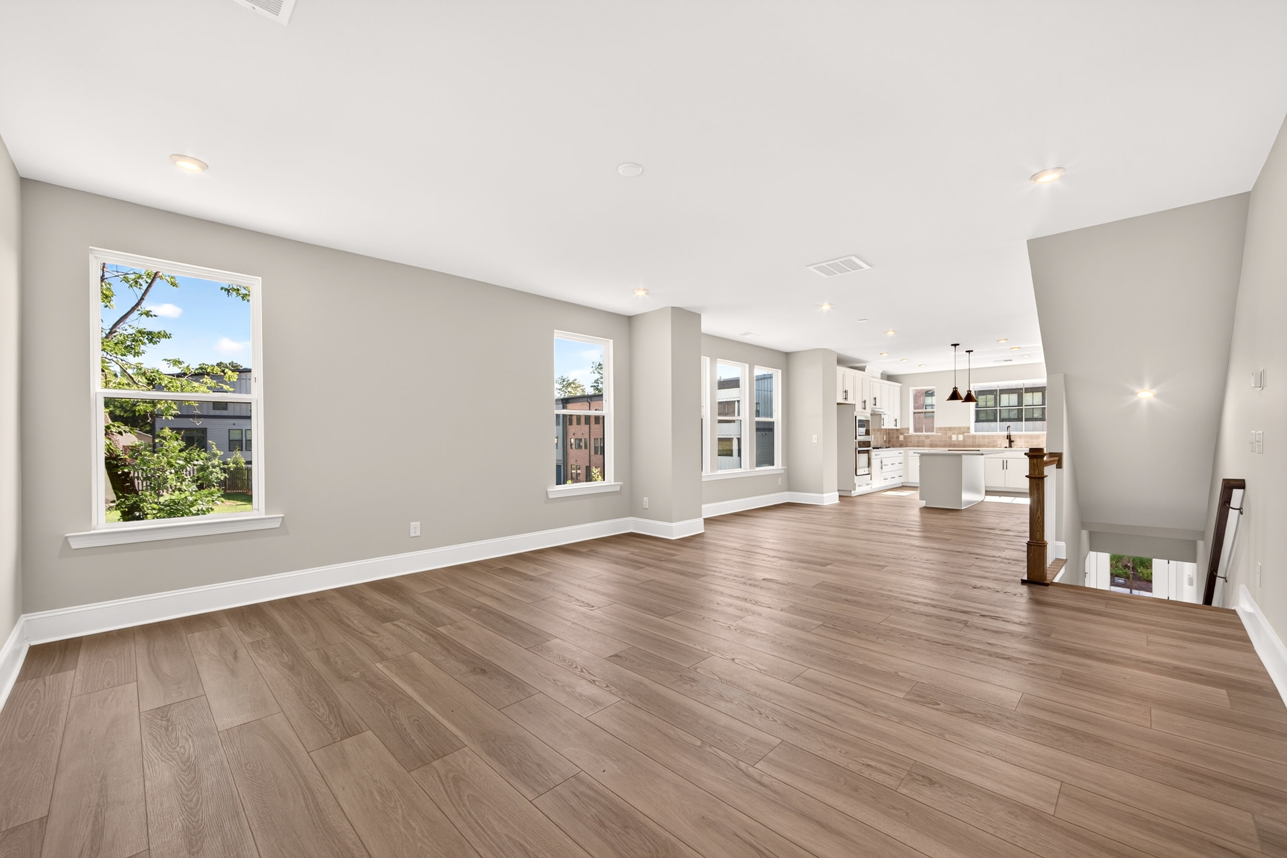 A spacious, modern living area with light wood floors in the foreground opens to a bright kitchen and staircase in the background.