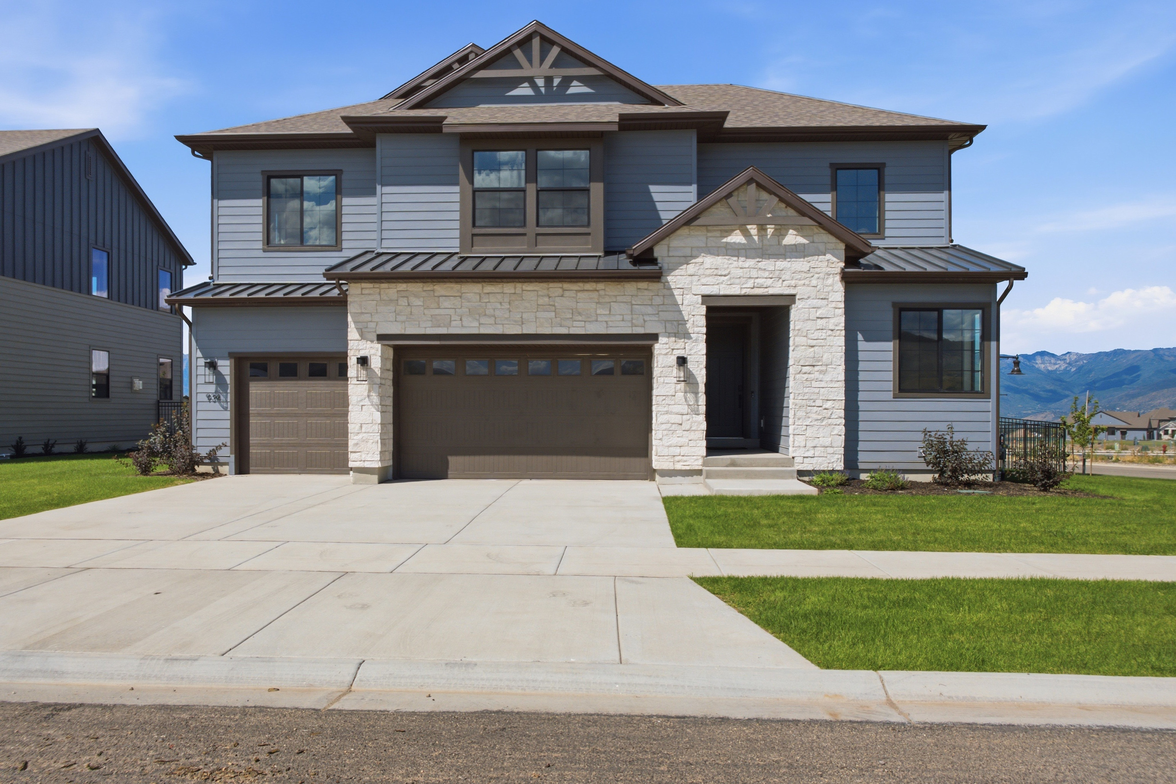 A modern two-story house with a light blue exterior and stone accents sits on a manicured lawn, with a paved driveway leading to its garages, all set against a backdrop of distant mountains under a clear blue sky.
